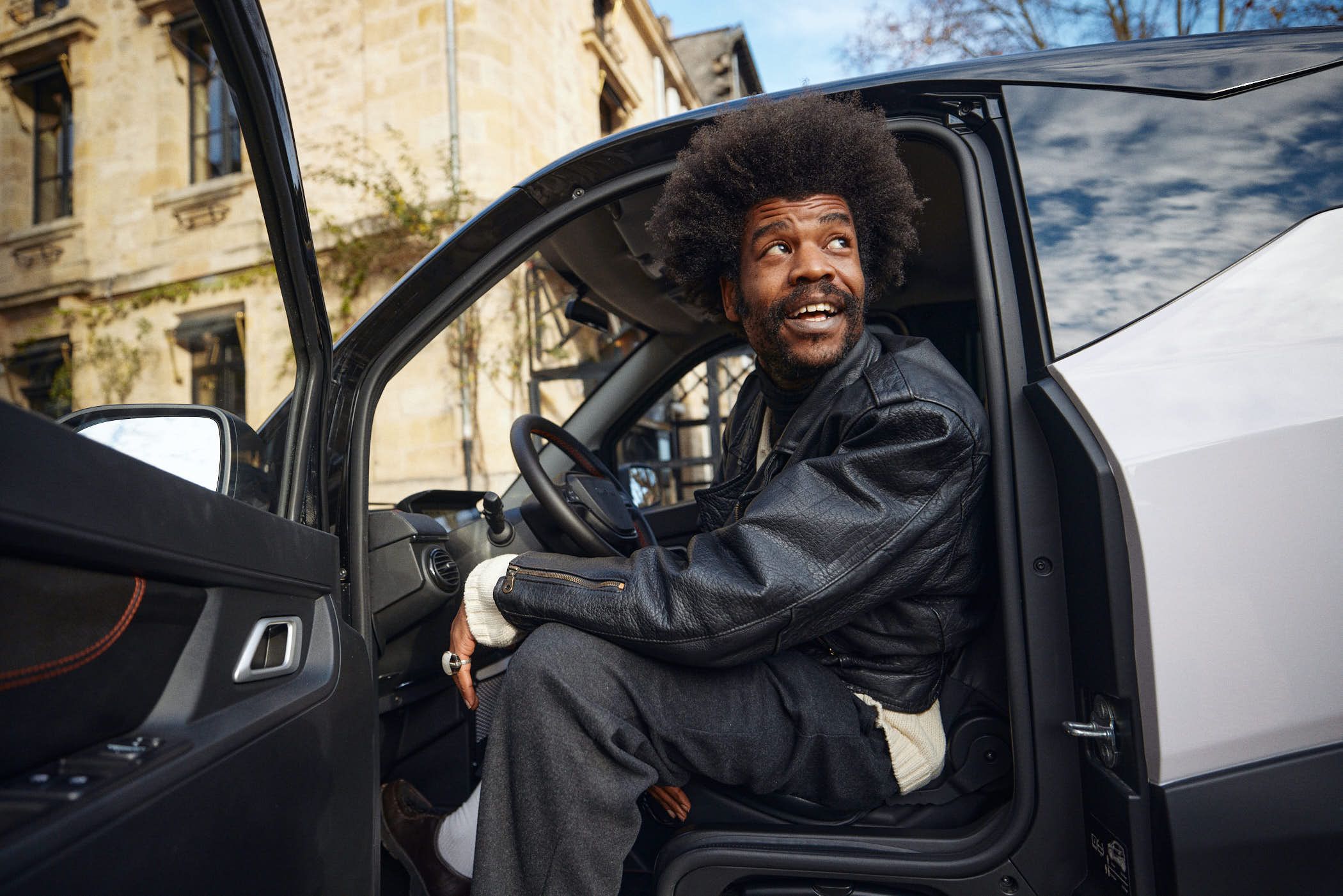 A man with a big afro sits in the driver's seat of a car, looking out the window with a serious expression. He is wearing a black leather jacket and dark pants. The background is.