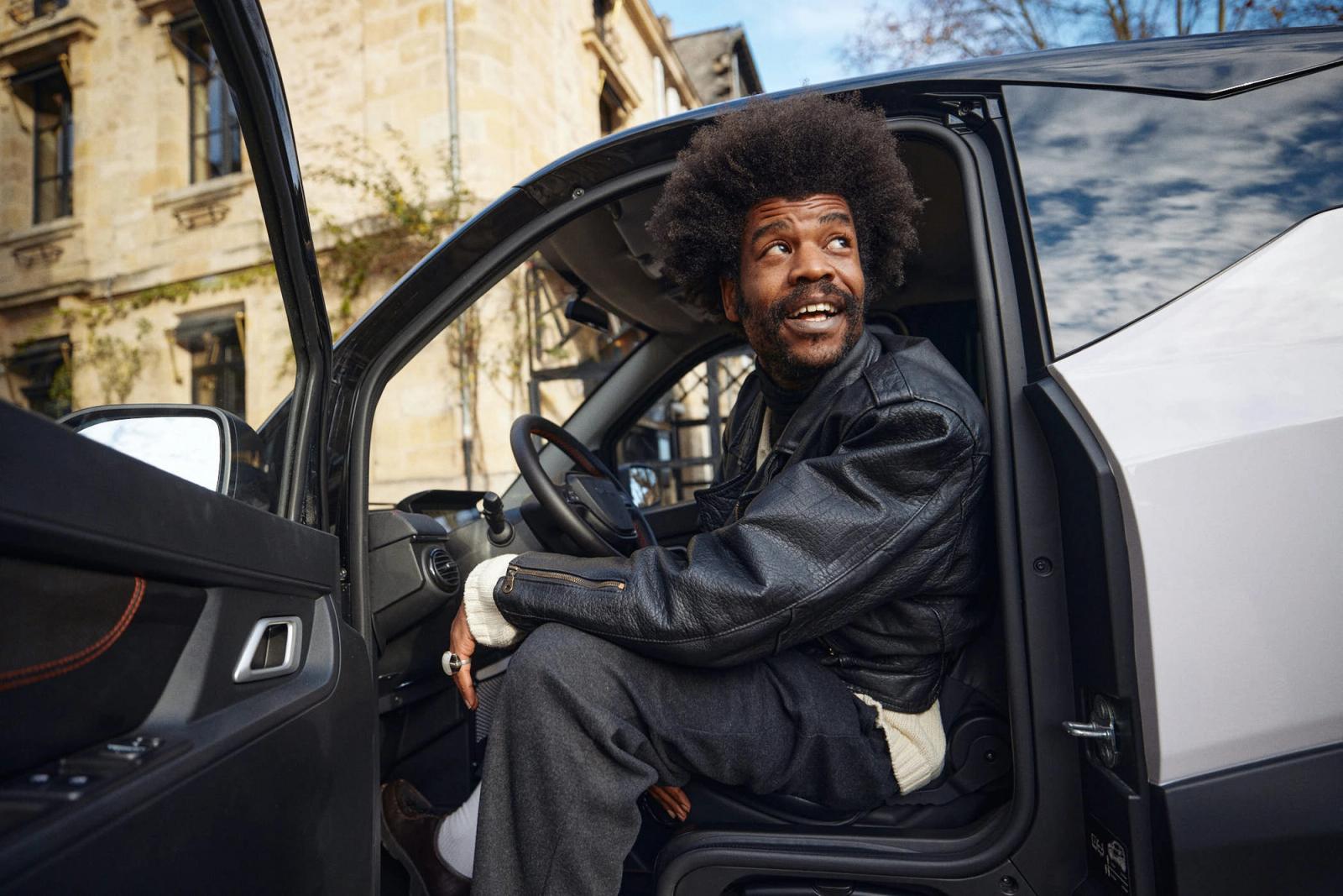 A man with a big afro sits in the driver's seat of a car, looking out the window with a serious expression. He is wearing a black leather jacket and dark pants. The background is.