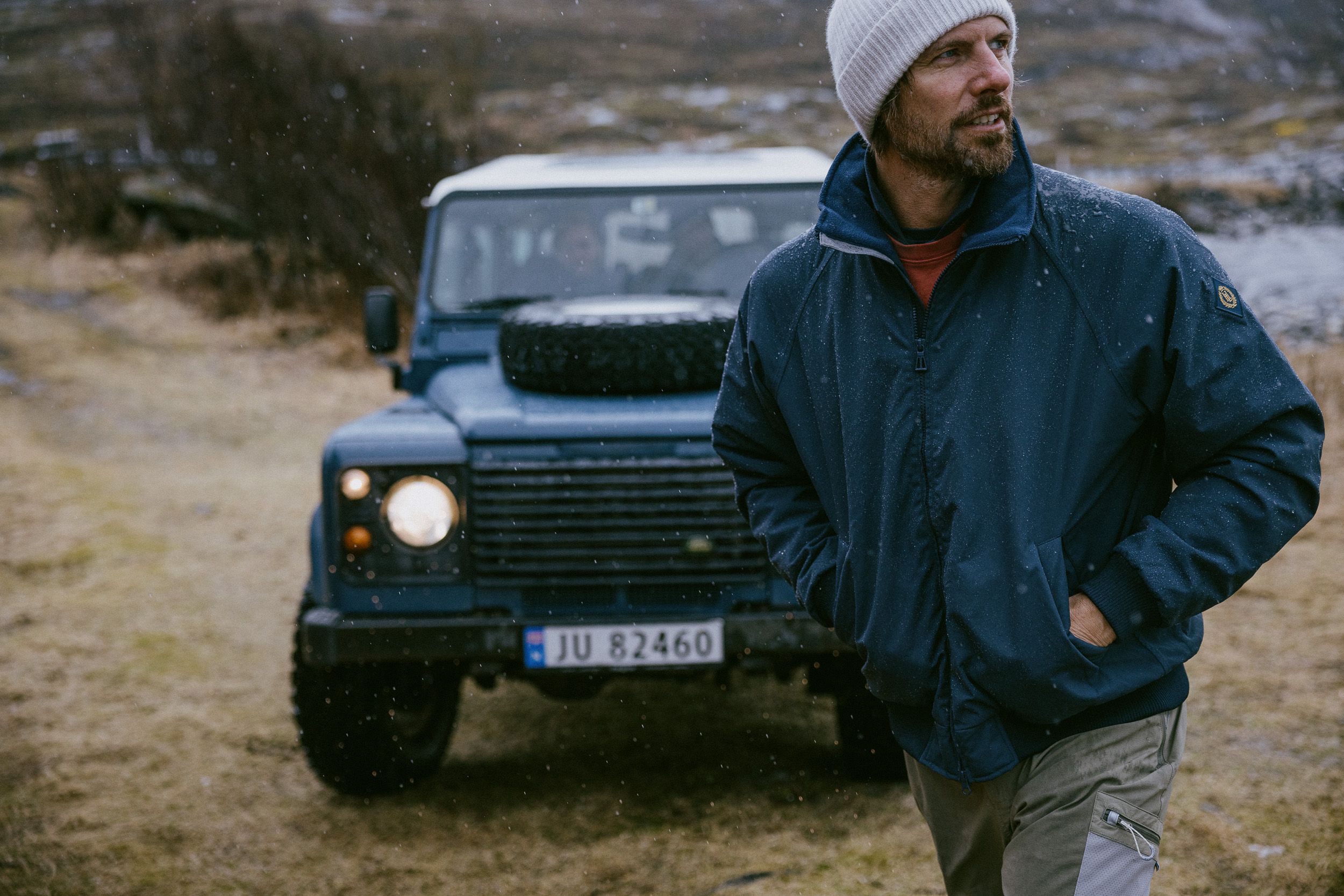 A man in a blue jacket and white beanie stands in front of a blue Land Rover Defender outdoors.