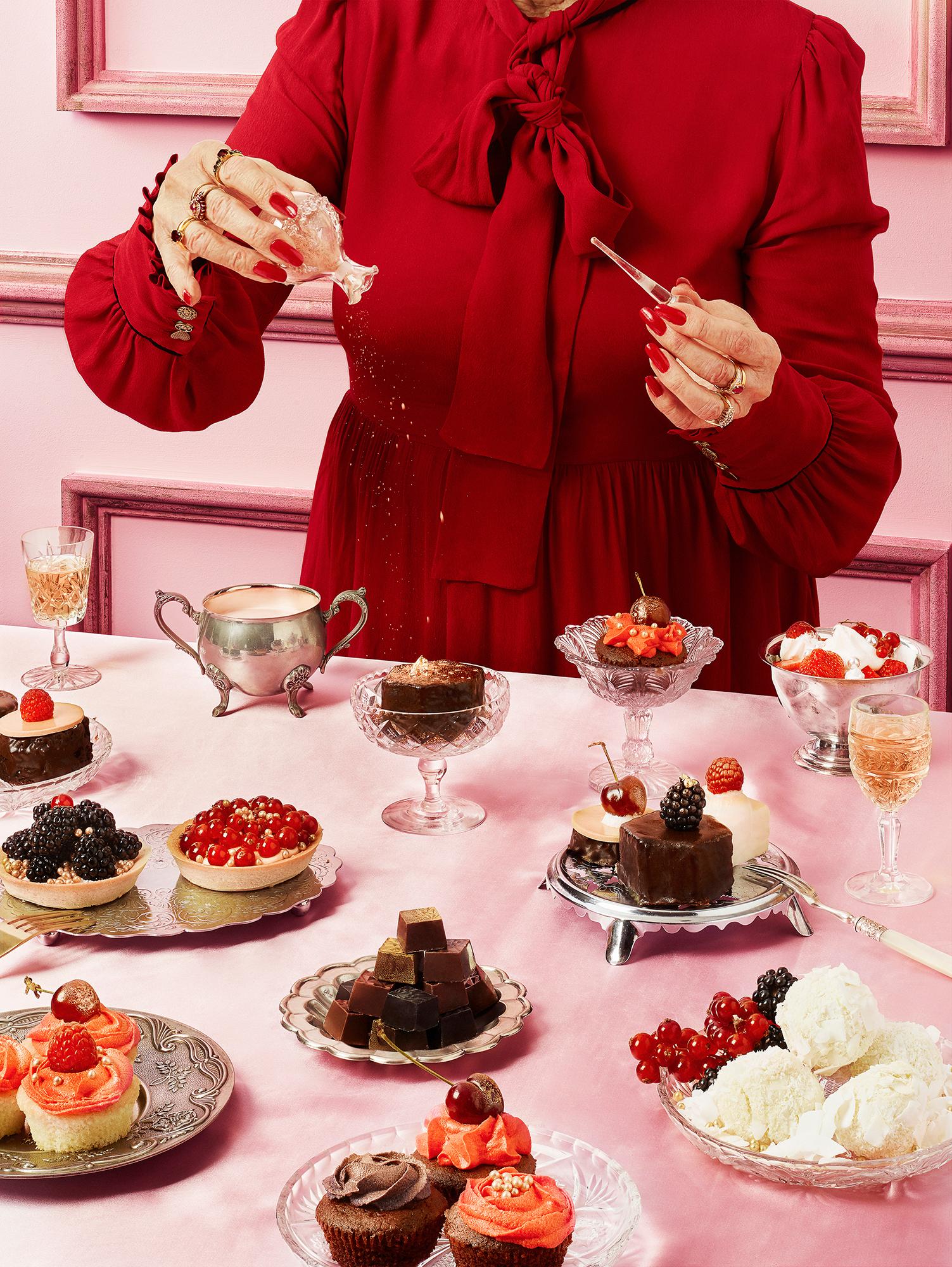 A woman in a red dress surrounded by cakes, cupcakes, and fruit.