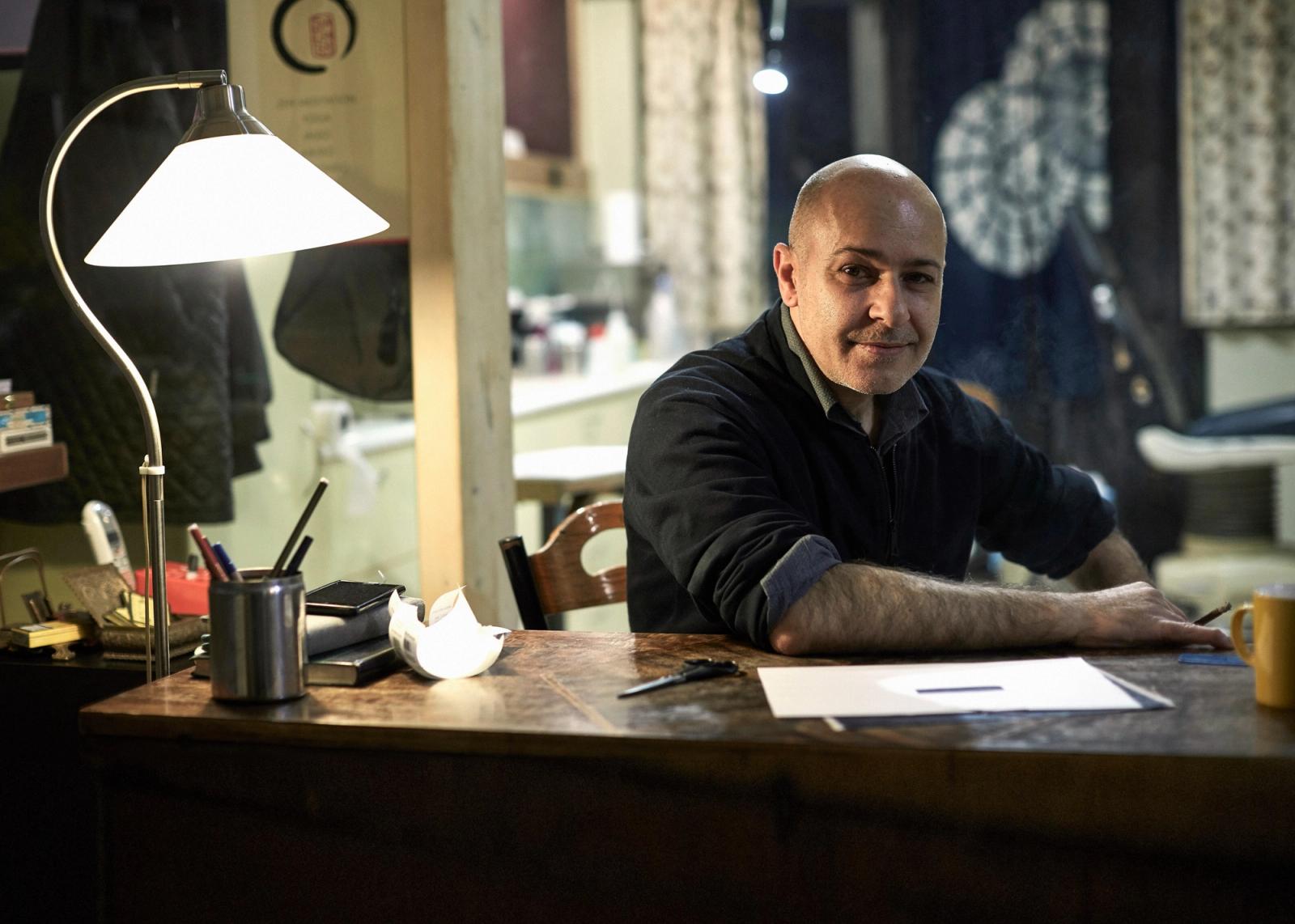 A man with a shaved head and a short, dark beard, wearing a black sweater and sitting at a desk with a lamp, papers, and a pen.