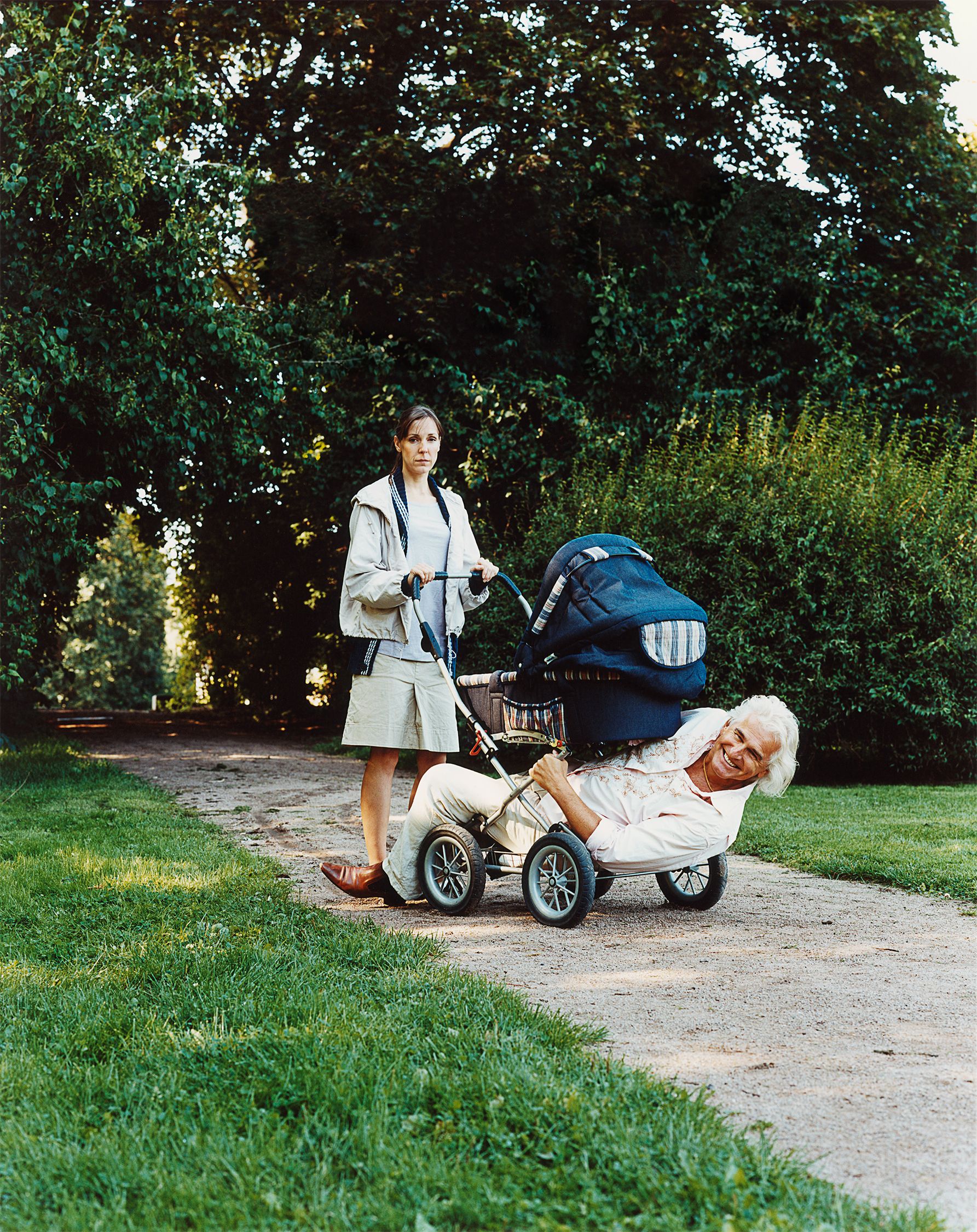 A woman in a beige coat and skirt pushing a stroller with a man in a white shirt and pants lying in it, on a dirt path in a park or garden.