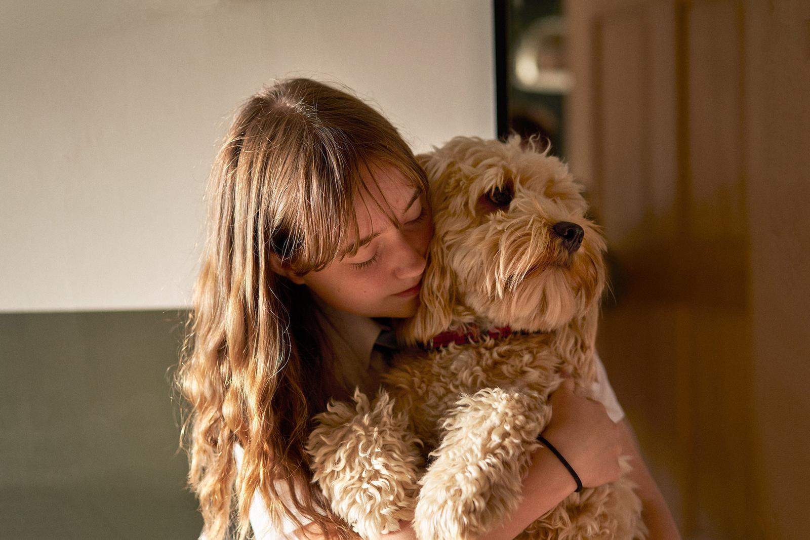 A young woman with long hair and a white shirt is hugging a small, fluffy, golden-brown dog.