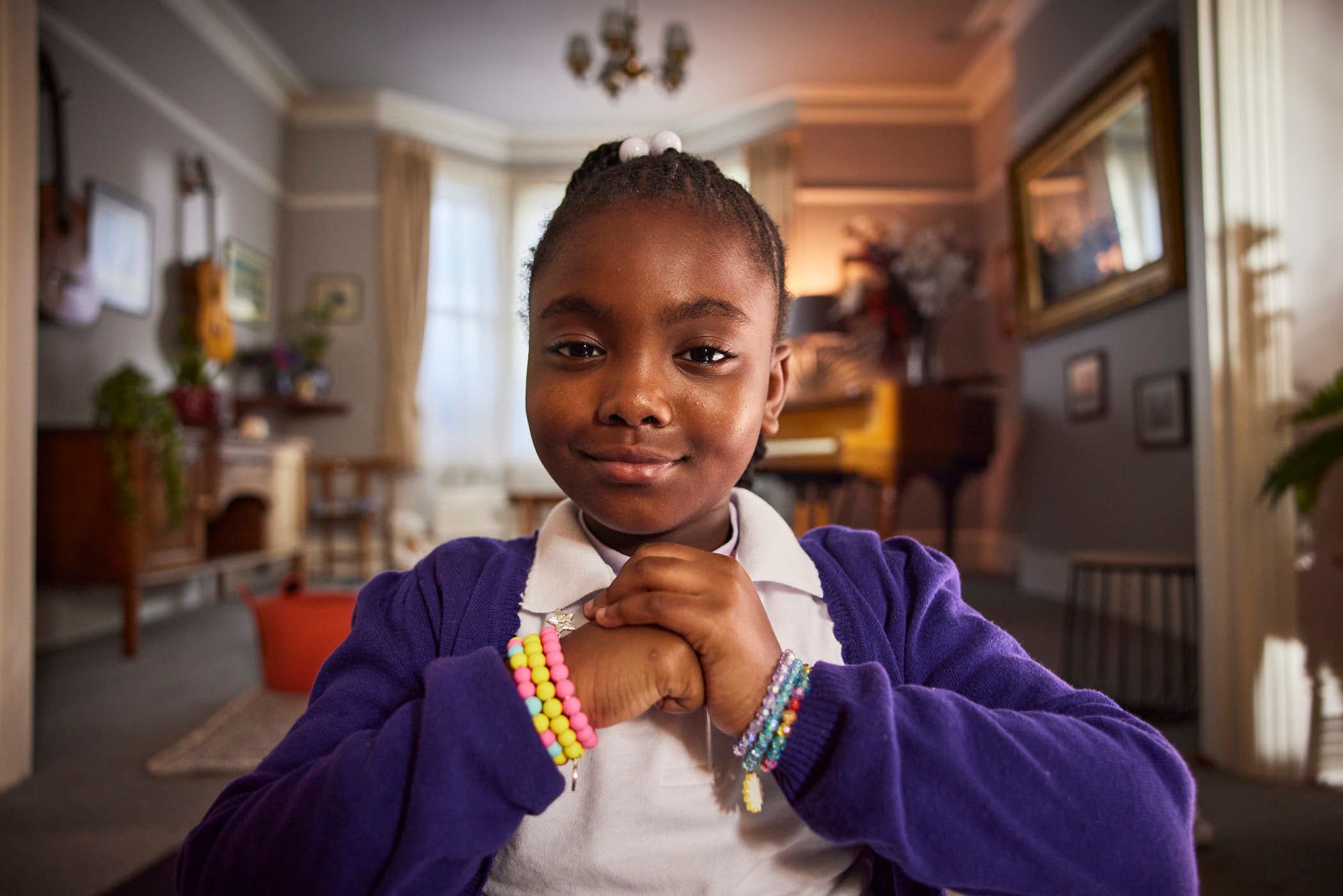 A young girl with her hands clasped together, wearing a purple cardigan over a white collared shirt and colorful beaded bracelets, smiles at the camera in a dimly lit, richly.