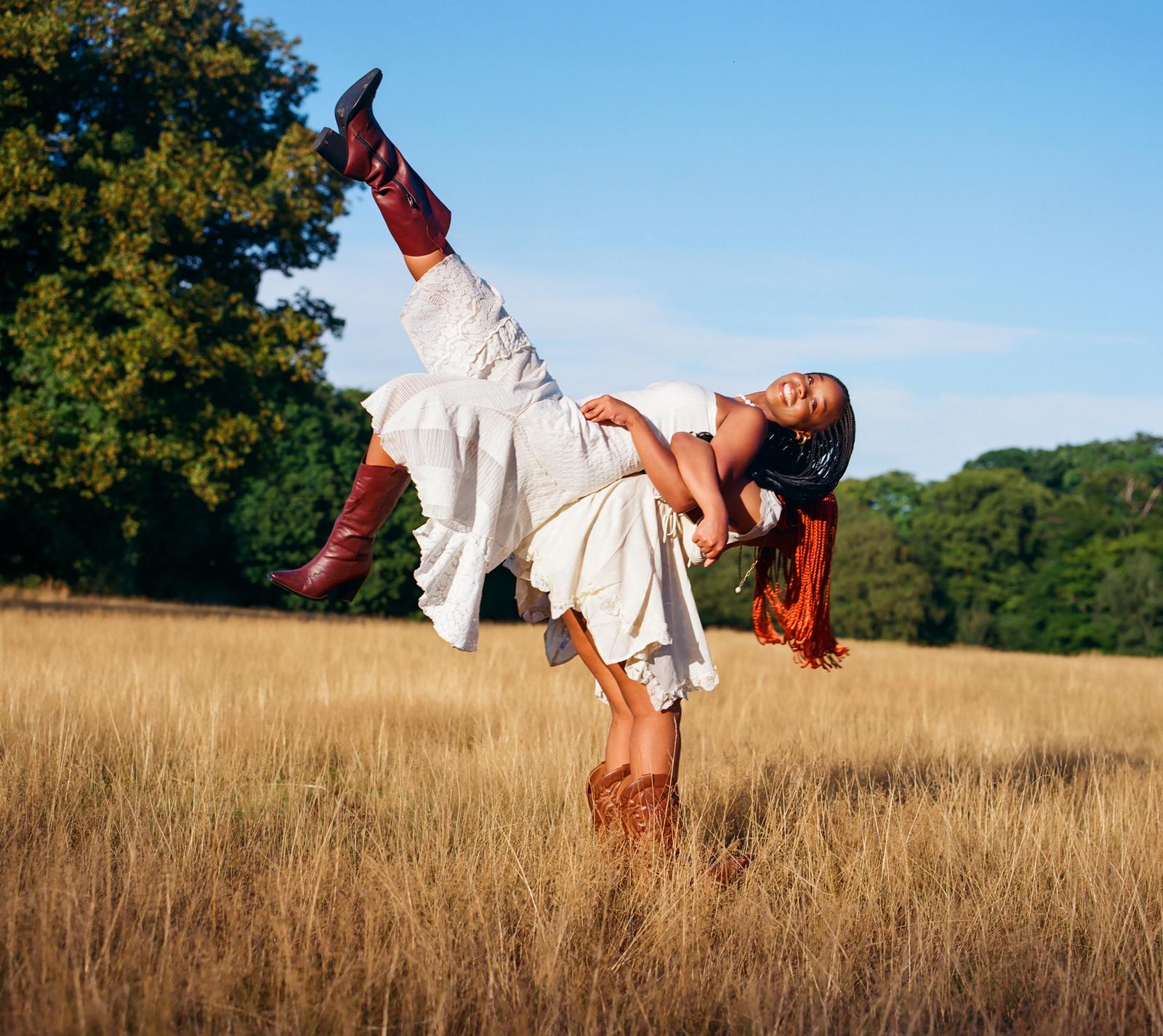 A woman in a white dress and brown boots is being lifted by another woman in a white dress and brown boots in a field of tall, golden grass.