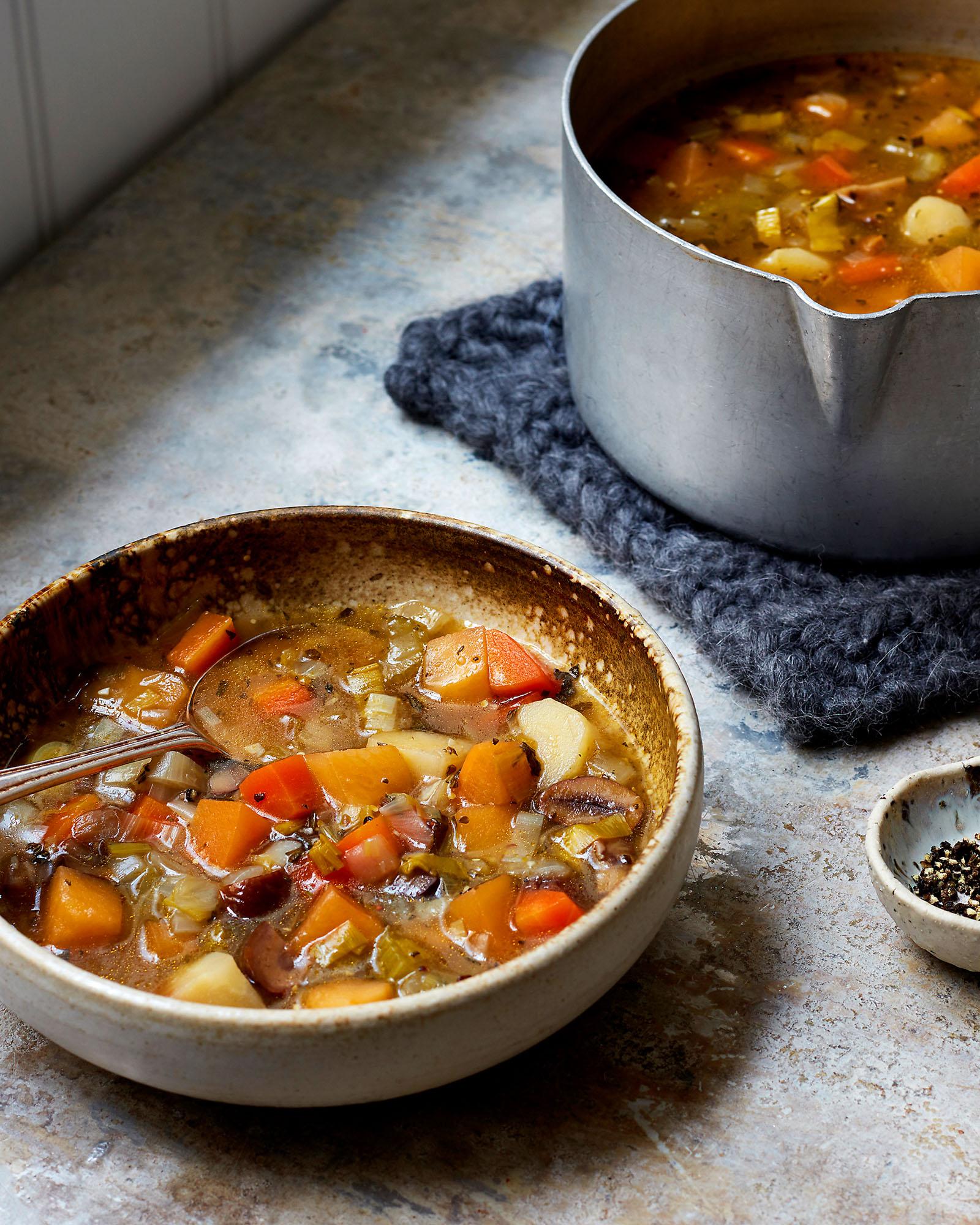 A bowl of vegetable soup with a spoon in it, with a pot of soup in the background.
