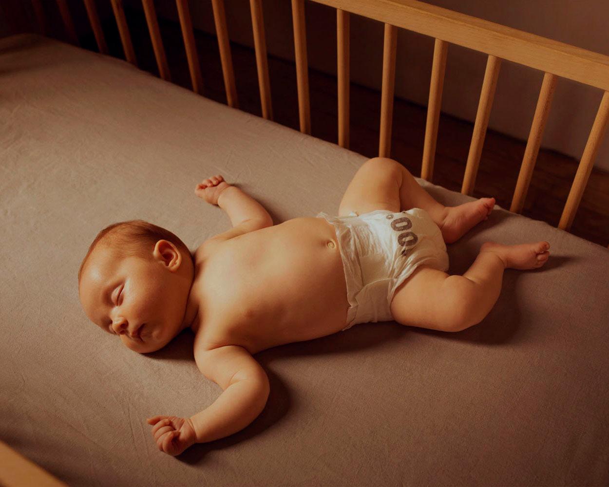 A sleeping infant in a diaper lies on a mattress in a crib.