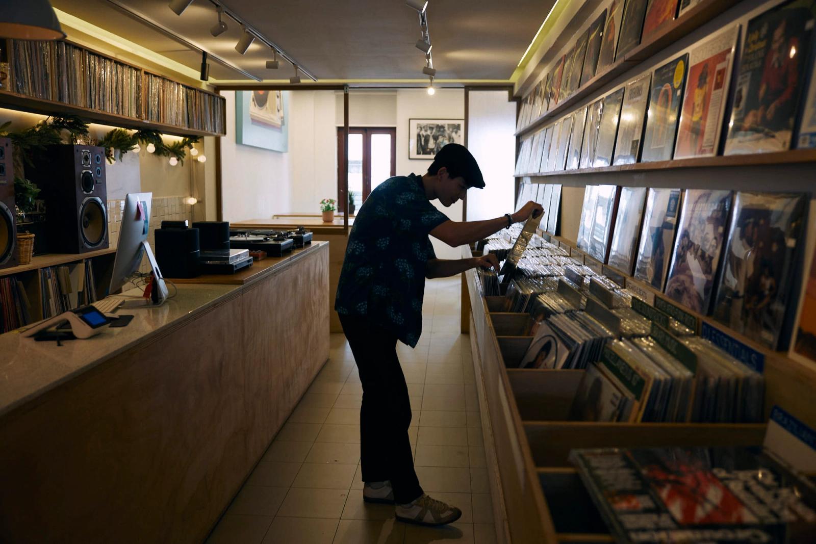 A man in a record store browses through vinyl records, wearing a blue Hawaiian shirt, black pants, and a black hat. The store's shelves are filled with records and a counter with.