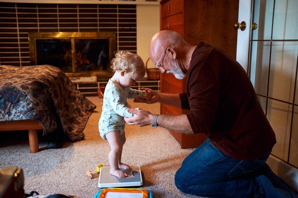 A grandfatherly man with a white beard and glasses kneels on the floor, gently guiding a toddler with blonde curls on a white scale. The child's eyes are fixed on the man's hands.