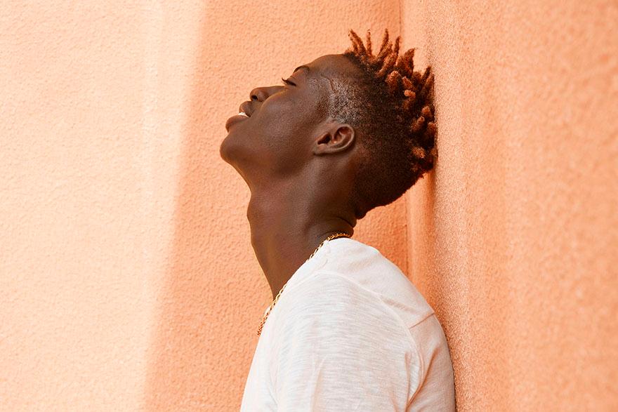 A young man with short, spiky hair and a white t-shirt leaning against a wall, looking up towards the sky.