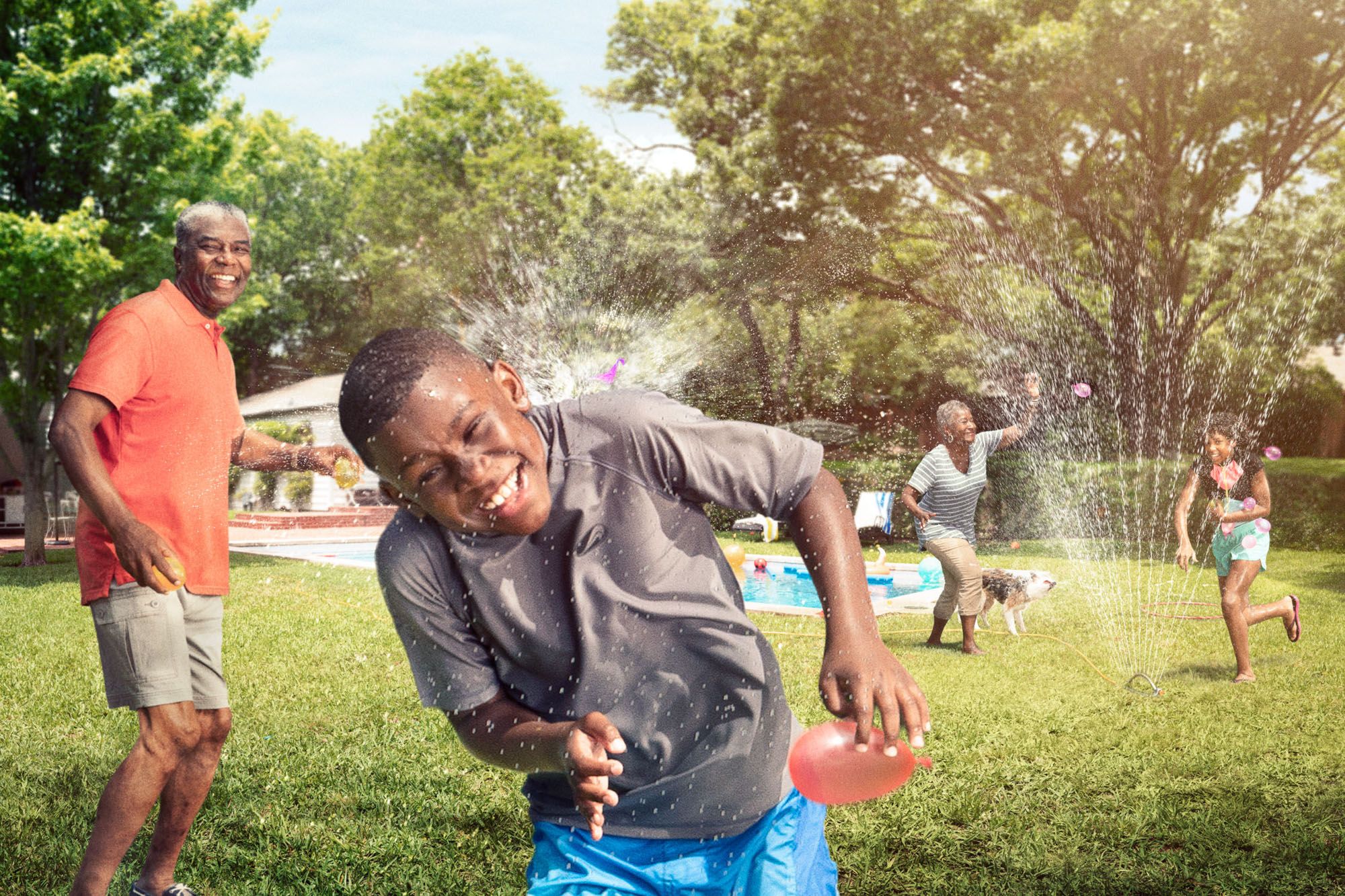 A group of people, including children and adults, are playing with water in a backyard setting, with a pool, trees, and a house in the background.