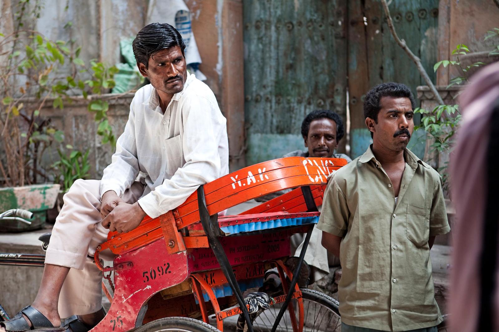 A man in a white shirt and beige pants sitting on a red rickshaw with two men in the background, one in a green shirt and one in a white shirt, in front of a weathered wooden door.