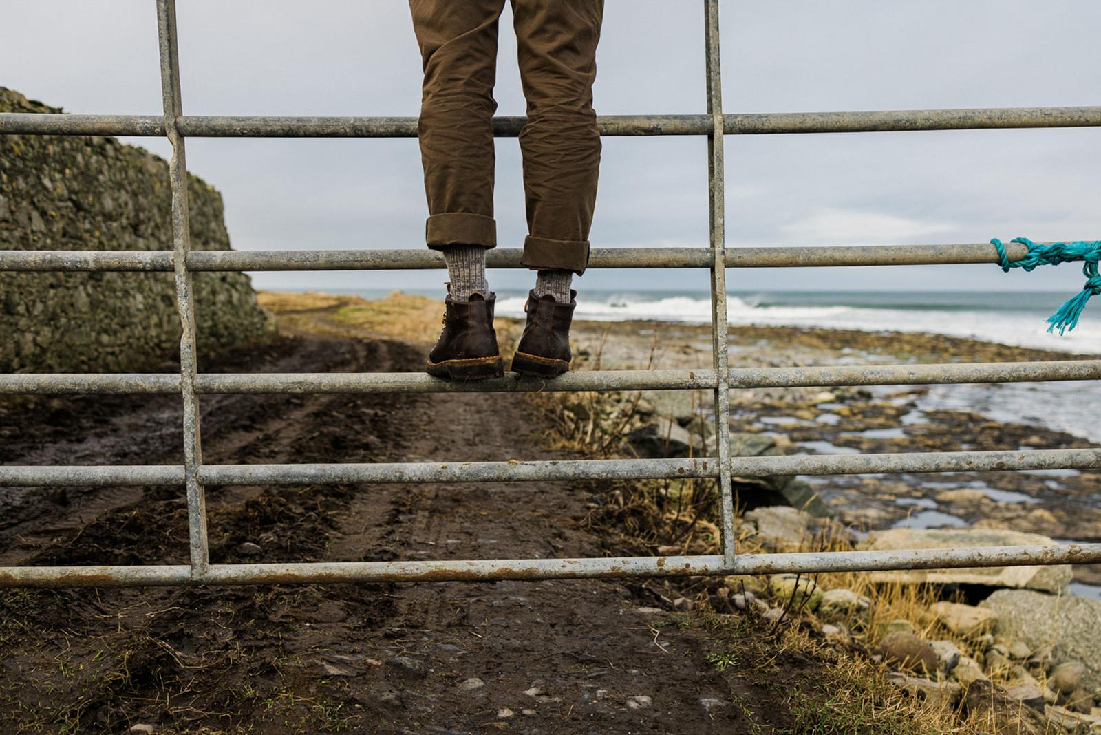 A person's legs are suspended from a metal gate overlooking a rugged coastline, with the person's feet dangling in the air.