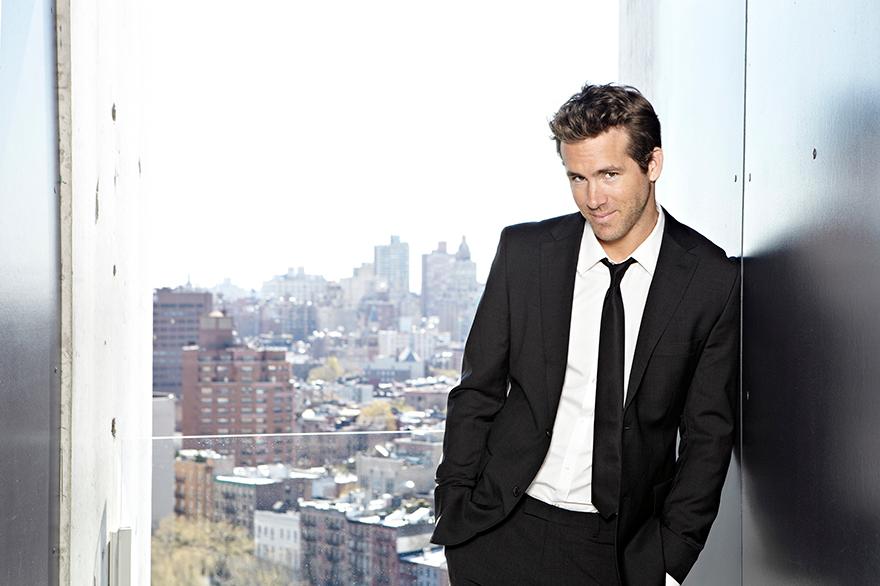 Actor Ryan Reynolds in a black suit, white shirt, and black tie, standing in front of a city skyline.