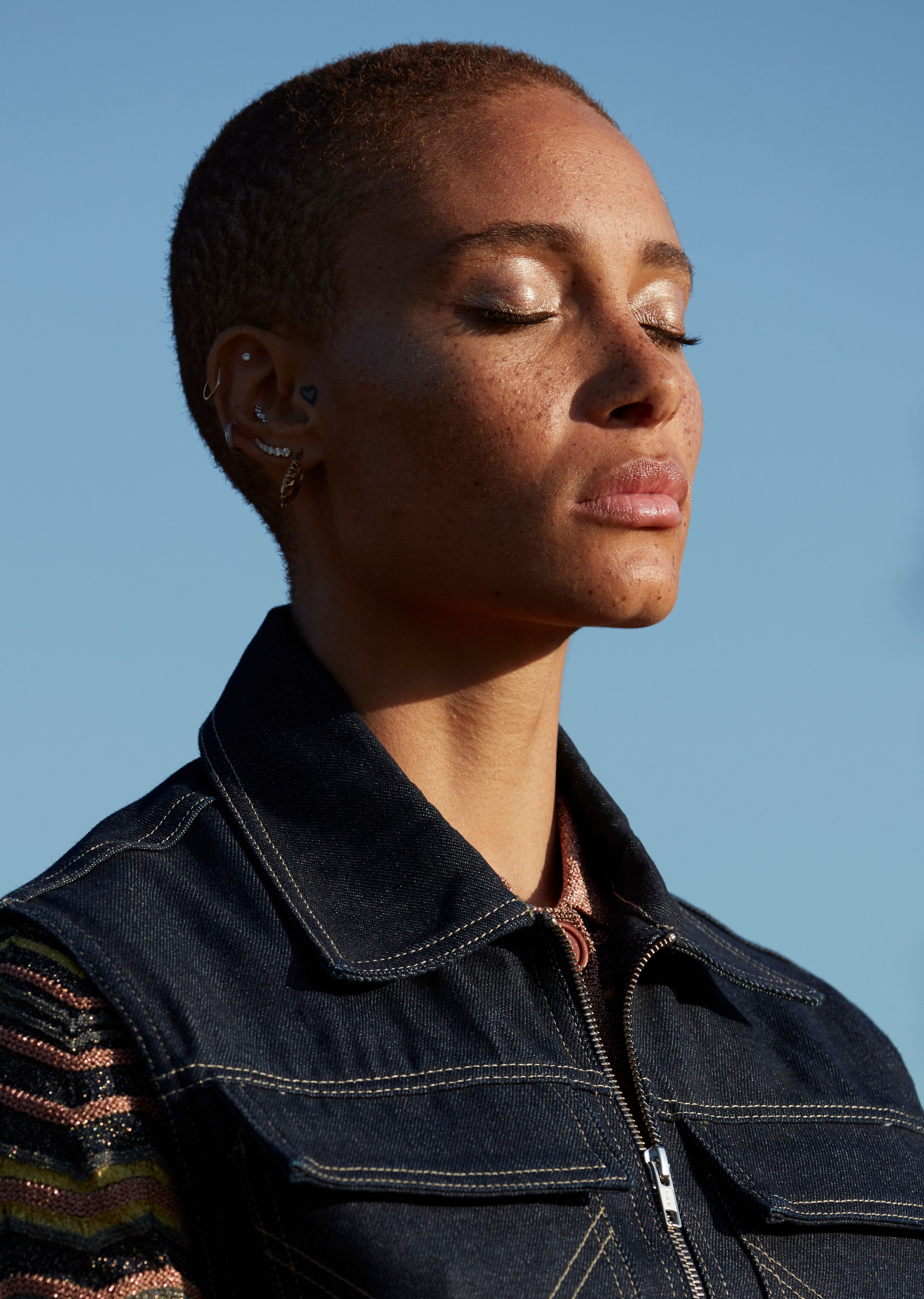 A woman with a shaved head and a black denim jacket with a zipper, standing in front of a blue sky.