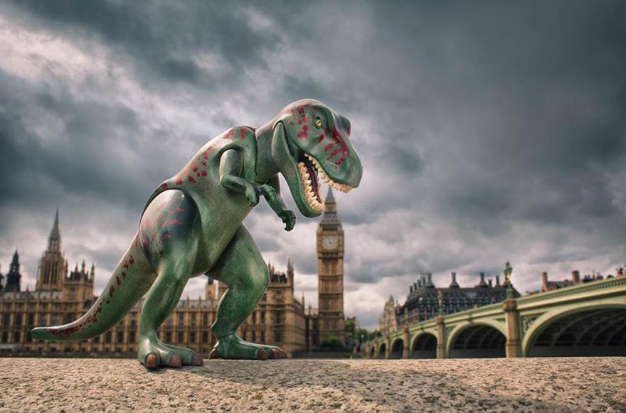 A green plastic toy T-Rex stands on a stone surface with a blurred view of London's iconic landmarks including Big Ben and Westminster Bridge in the background.