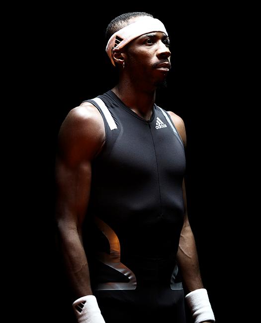 A man wearing a black and white Adidas sports suit, with a white headband and a serious expression, standing in a darkened room.