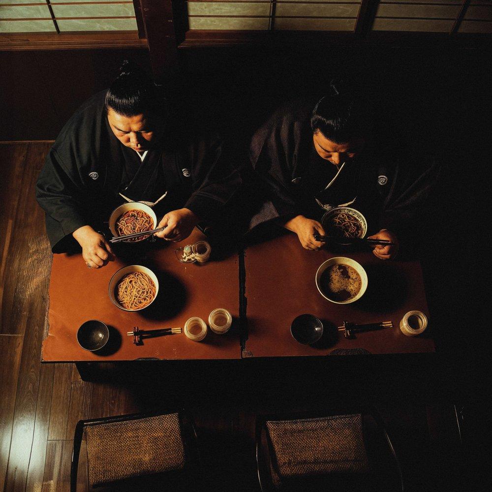 Two people sitting at a table eating noodles with chopsticks.
