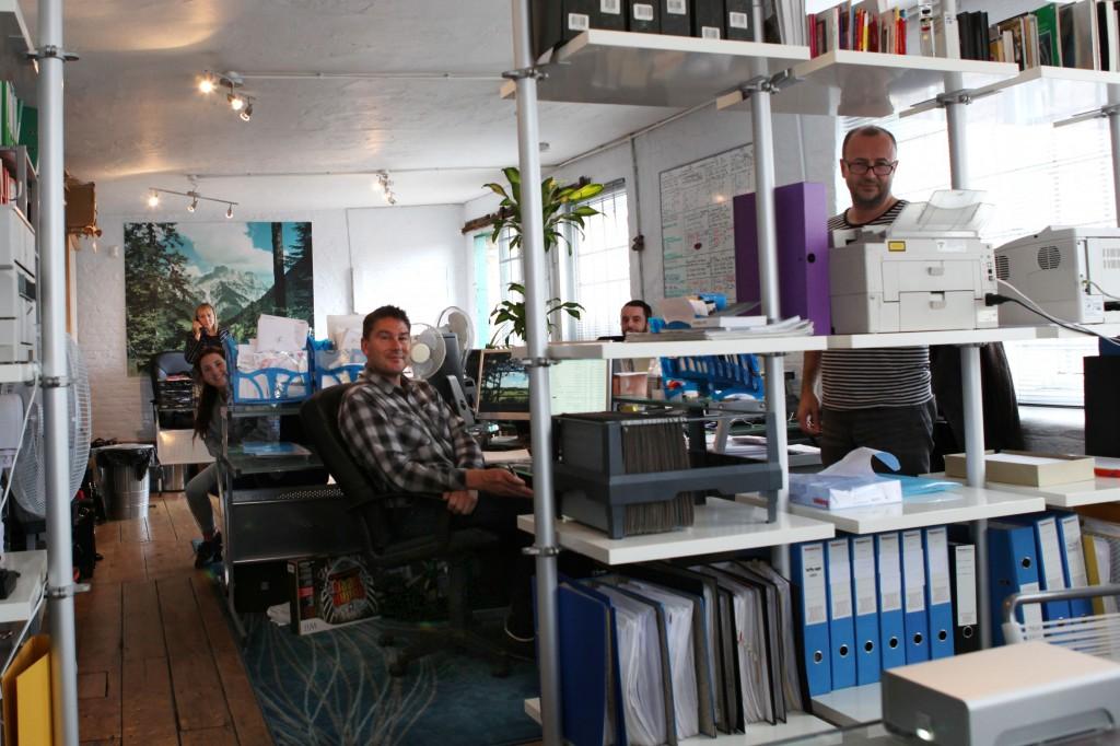 A group of people sitting at desks in a large, open office space with white walls and wooden floors, surrounded by rows of computer monitors and office equipment.