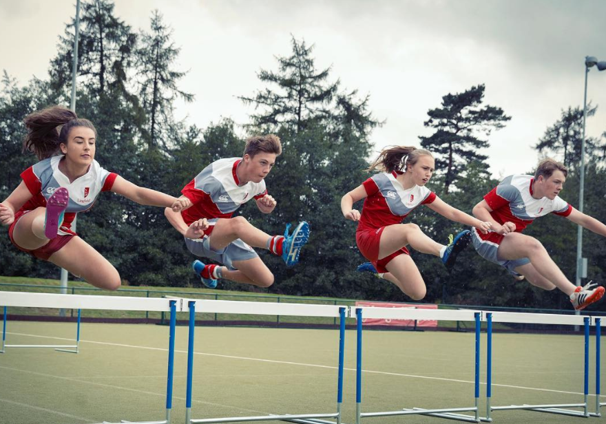 A group of children in red and white sports uniforms are running in a stadium, jumping over hurdles.