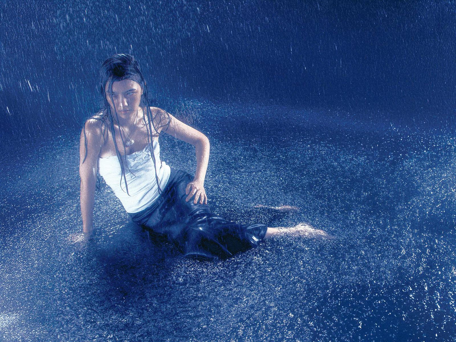 A woman in a white tank top and black pants is sitting in a pool of water, surrounded by water droplets.
