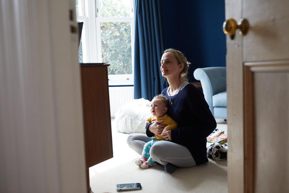 A woman sits on the floor with a baby in her lap, both looking up at something out of frame. The woman is smiling, but the baby's expression is neutral.