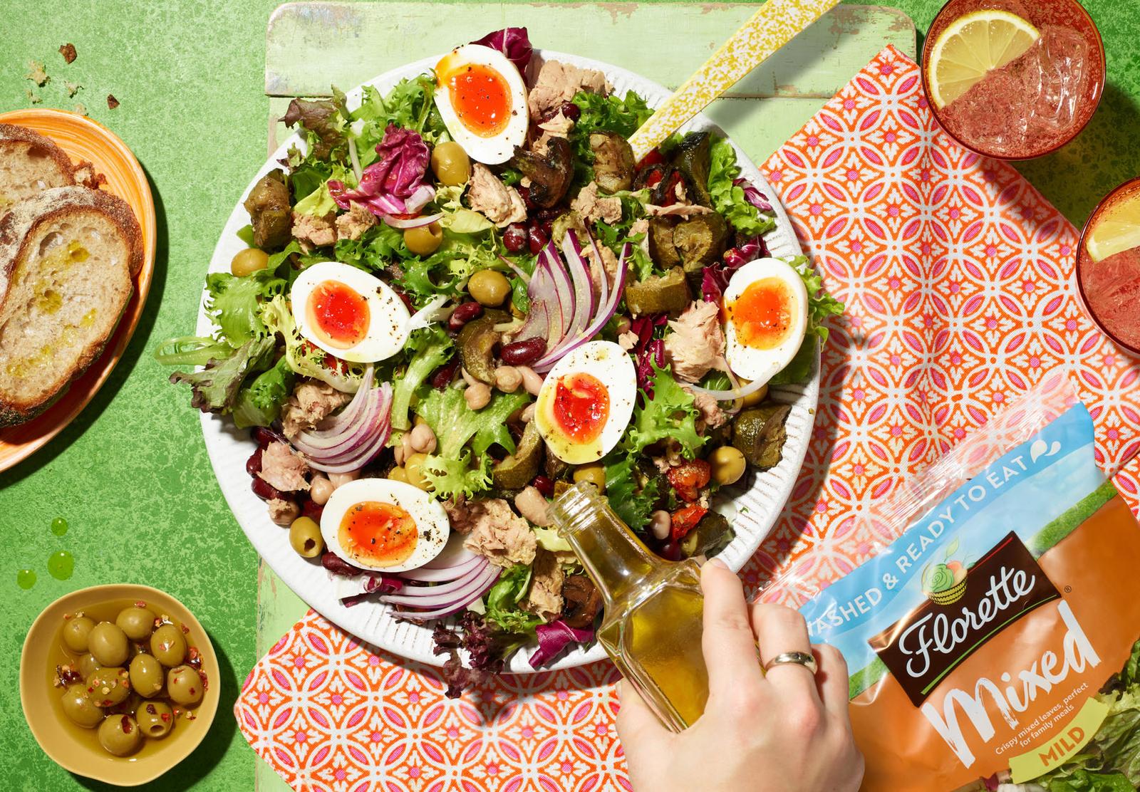 A salad with mixed greens, cherry tomatoes, avocado, and a fried egg, served on a white plate, with a bottle of olive oil and a slice of bread on the side.