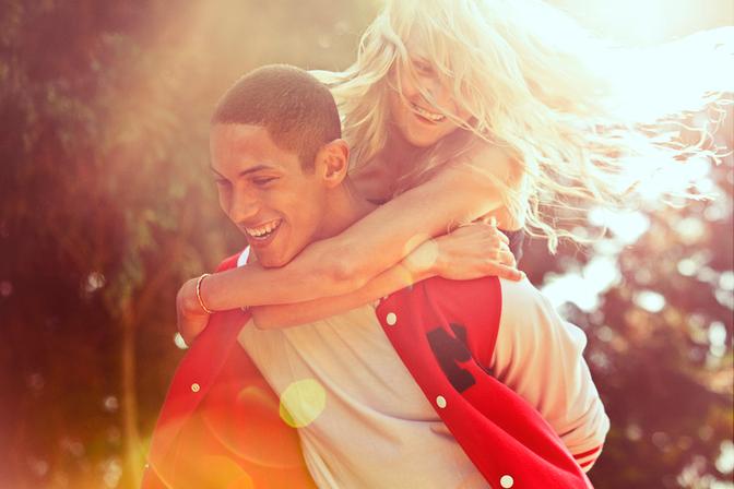 A young woman with long blonde hair is being lifted onto the shoulders of a young man with a red and white striped rugby jersey, both smiling and laughing.