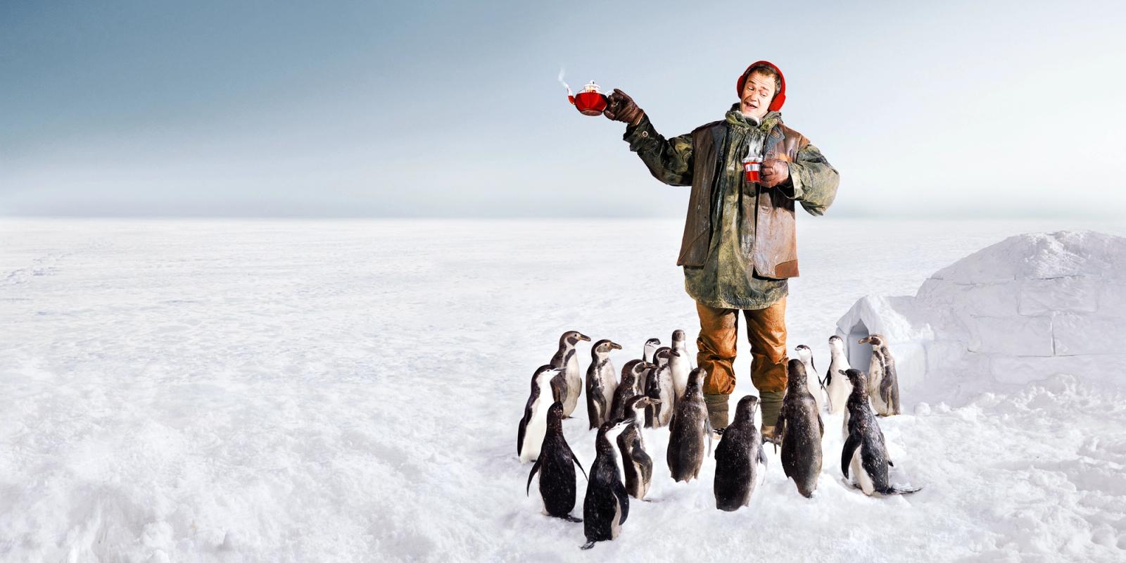 A man in a wetsuit and a red hat is standing on a snow-covered ice field, surrounded by penguins, holding a cup of hot coffee.