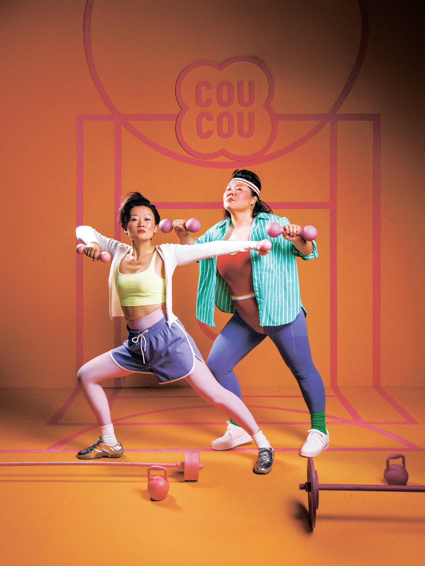 Two women in 1980s-style workout clothes lifting dumbbells in a gym.