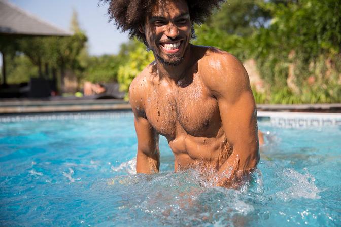 A shirtless man with a beard and a big smile, standing in a swimming pool, surrounded by water splashes.