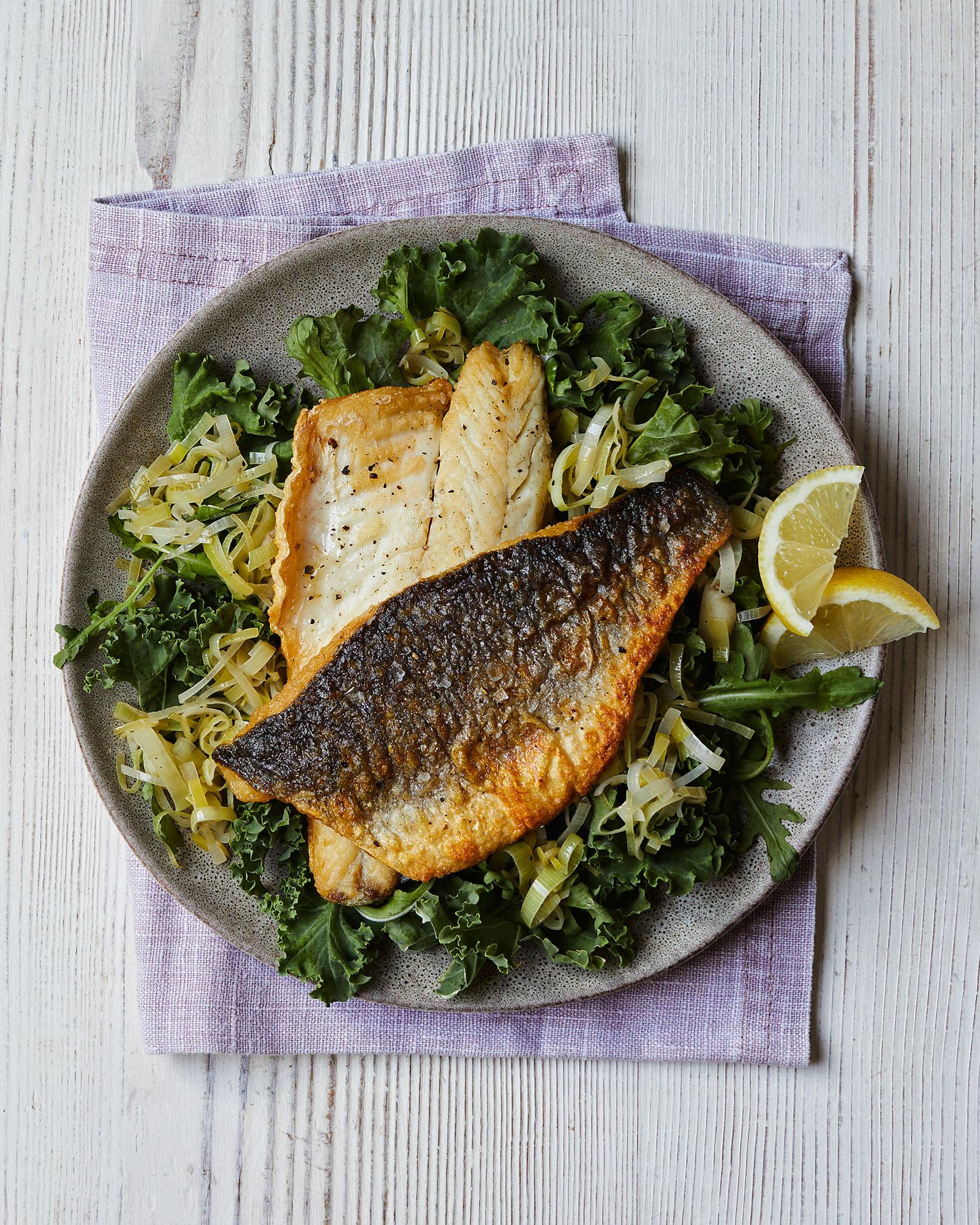A plate features pan-seared fish, greens, lemon slice, and herbs.