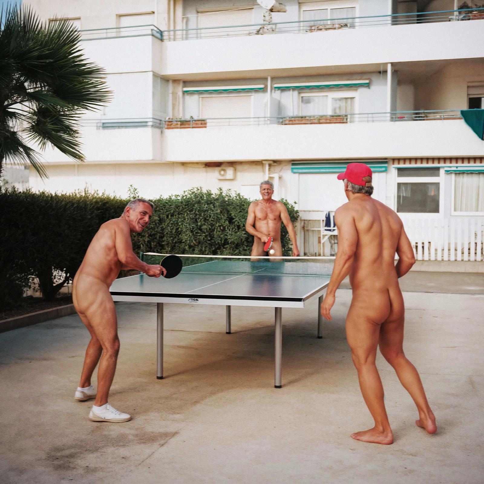 Three nude men playing table tennis in a courtyard, one wearing a red baseball cap.