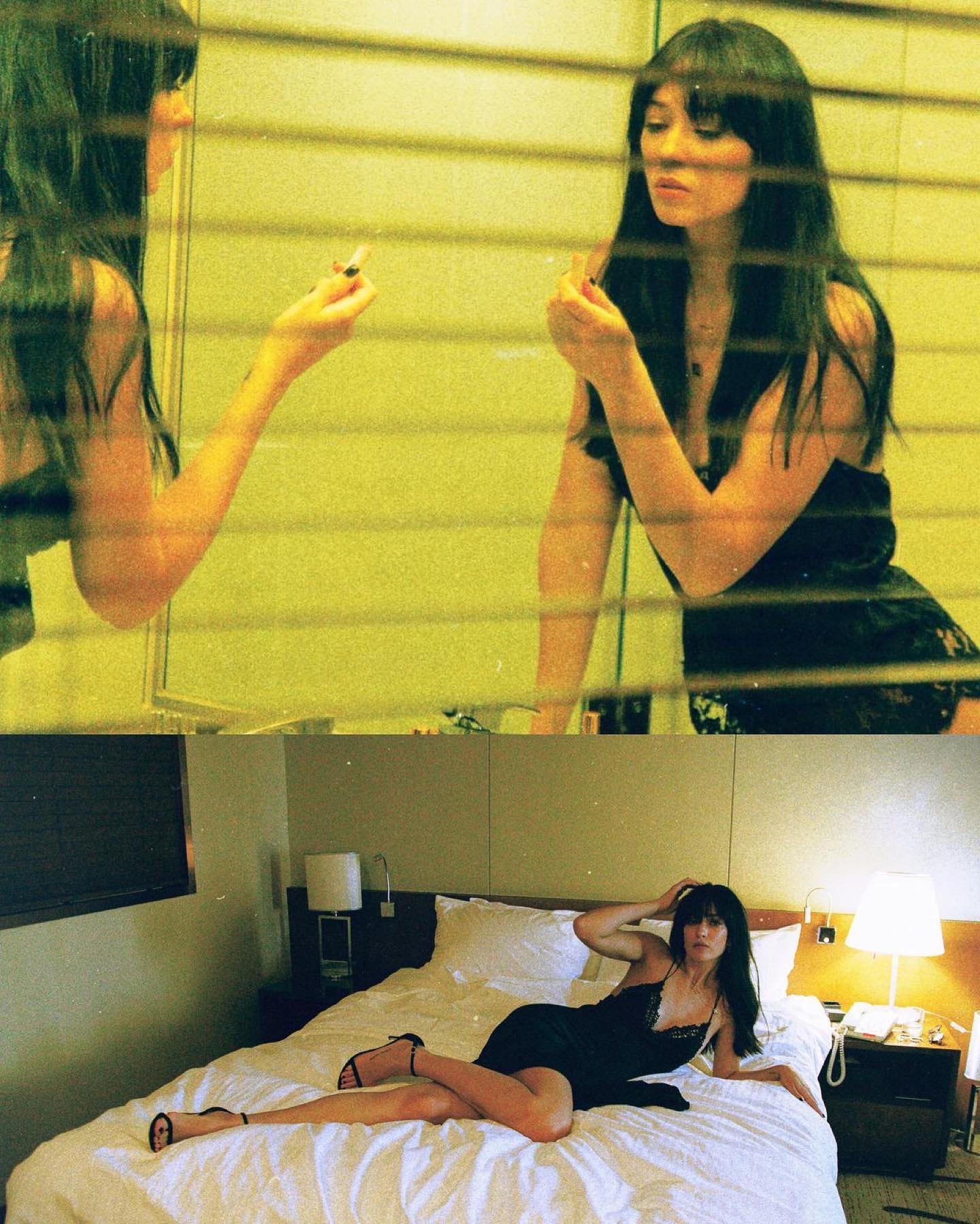 Darina Umka sitting on a bed, wearing a black dress and high heels, looking at her reflection in a mirror.