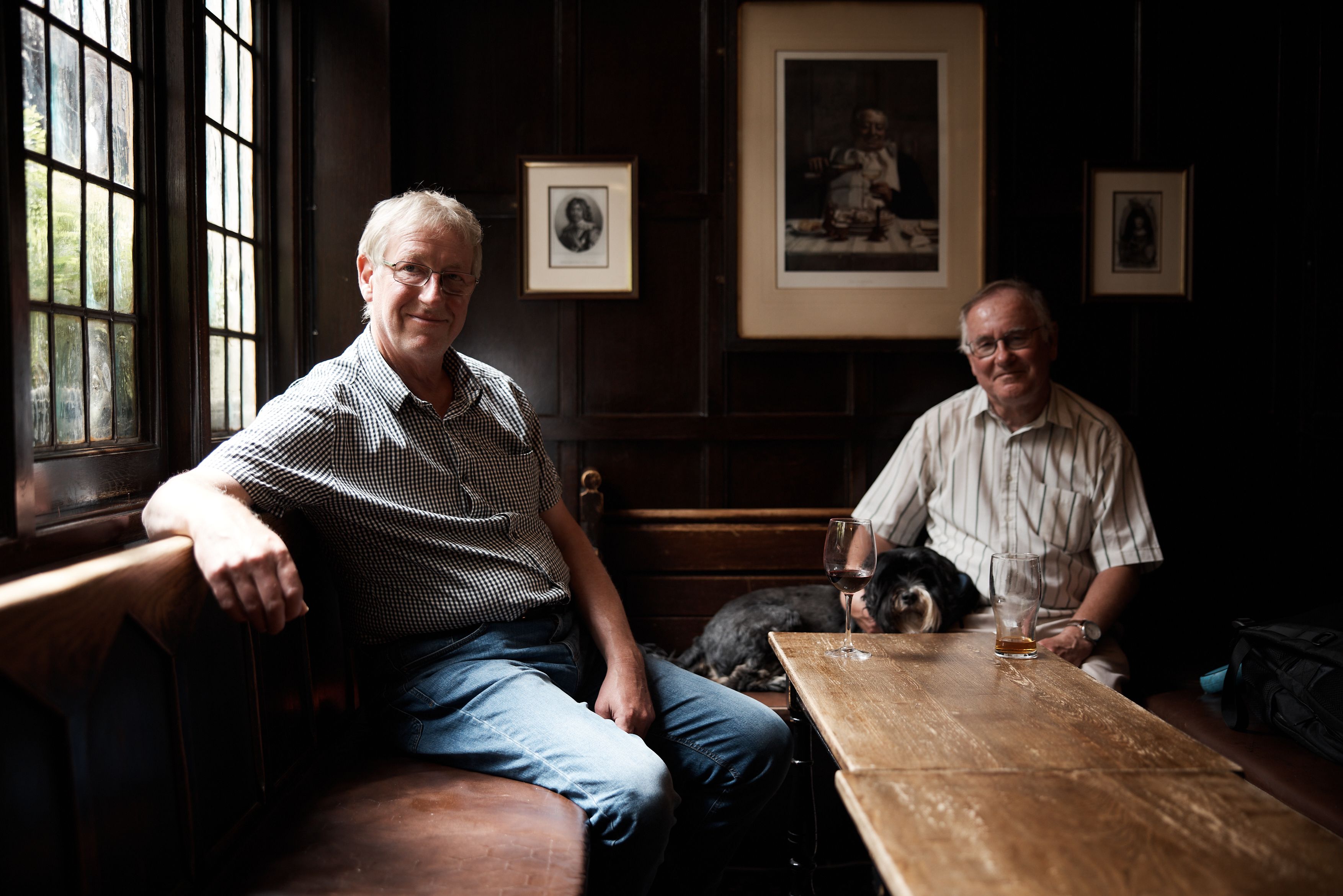 Two older men with drinks in a pub, one accompanied by a dog.