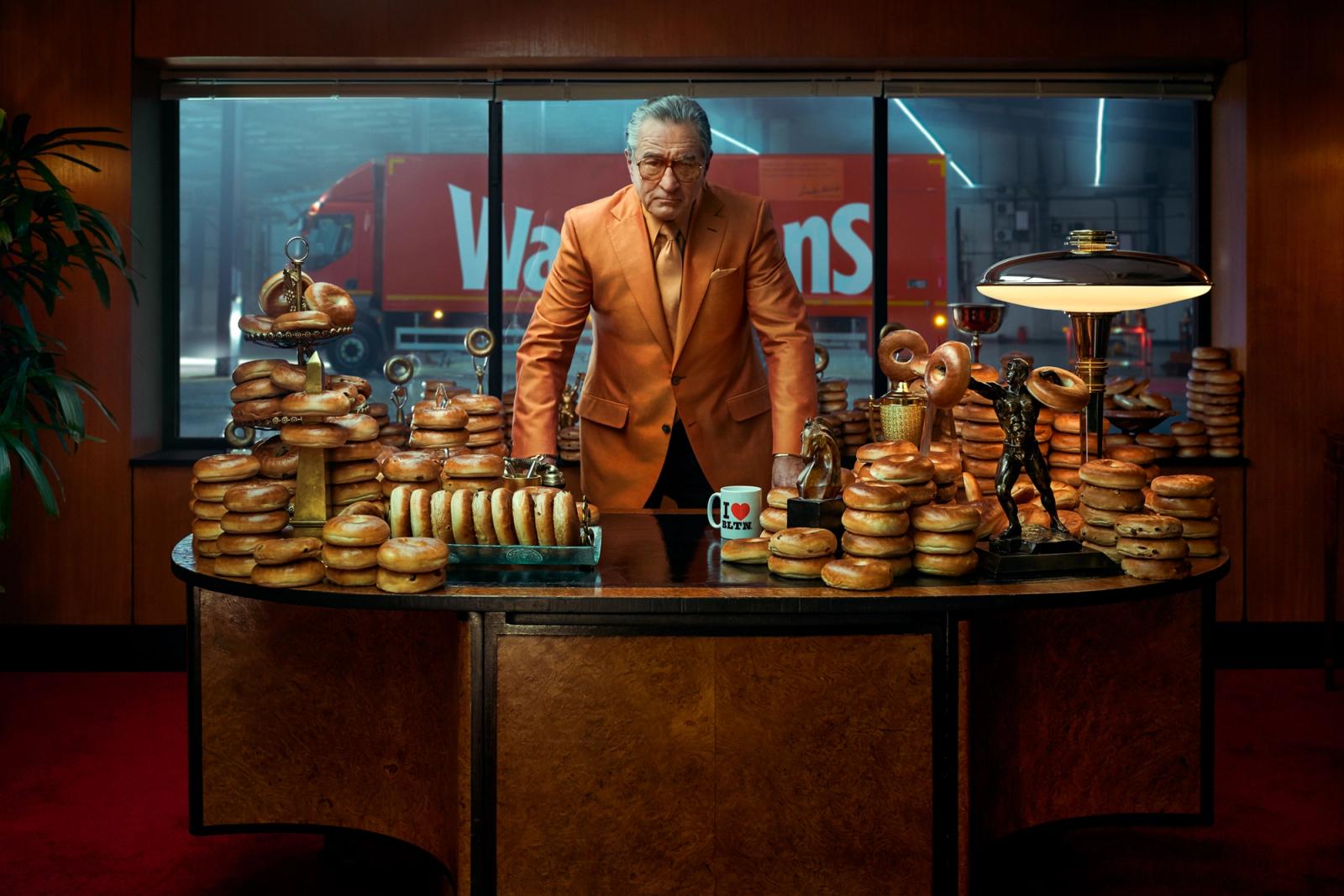 A man in a bright orange suit standing behind a large desk covered in stacks of bagels, with a red truck visible through the window behind him.
