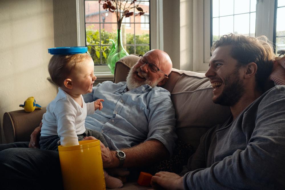 A young child with a blue container on their head sits on a couch with an older man and a younger man, all smiling at each other. The older man has a white beard and is wearing a.