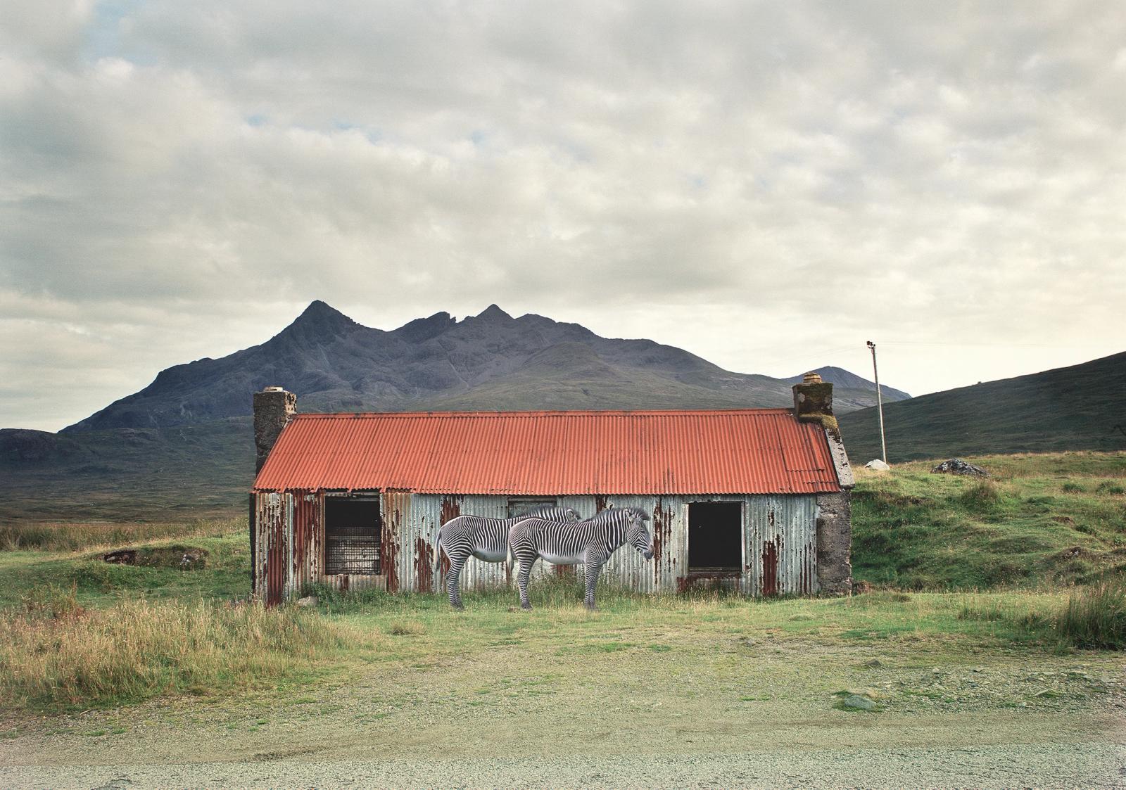 A photograph of a small, old, and abandoned house in a rural landscape, with a mountain in the background.