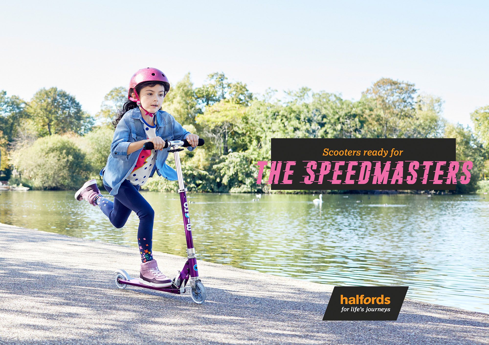 A young girl on a scooter, wearing a pink helmet, riding on a path near a lake.