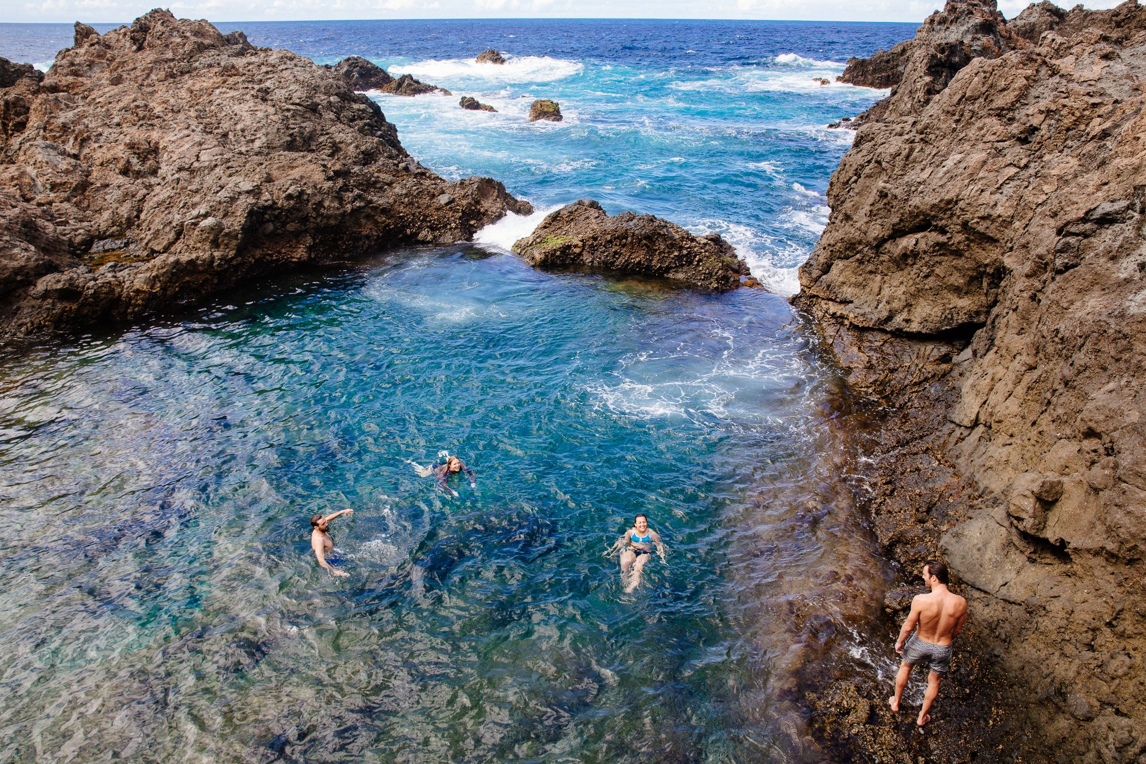 A group of people swimming in a natural rock pool surrounded by large boulders and ocean waves.