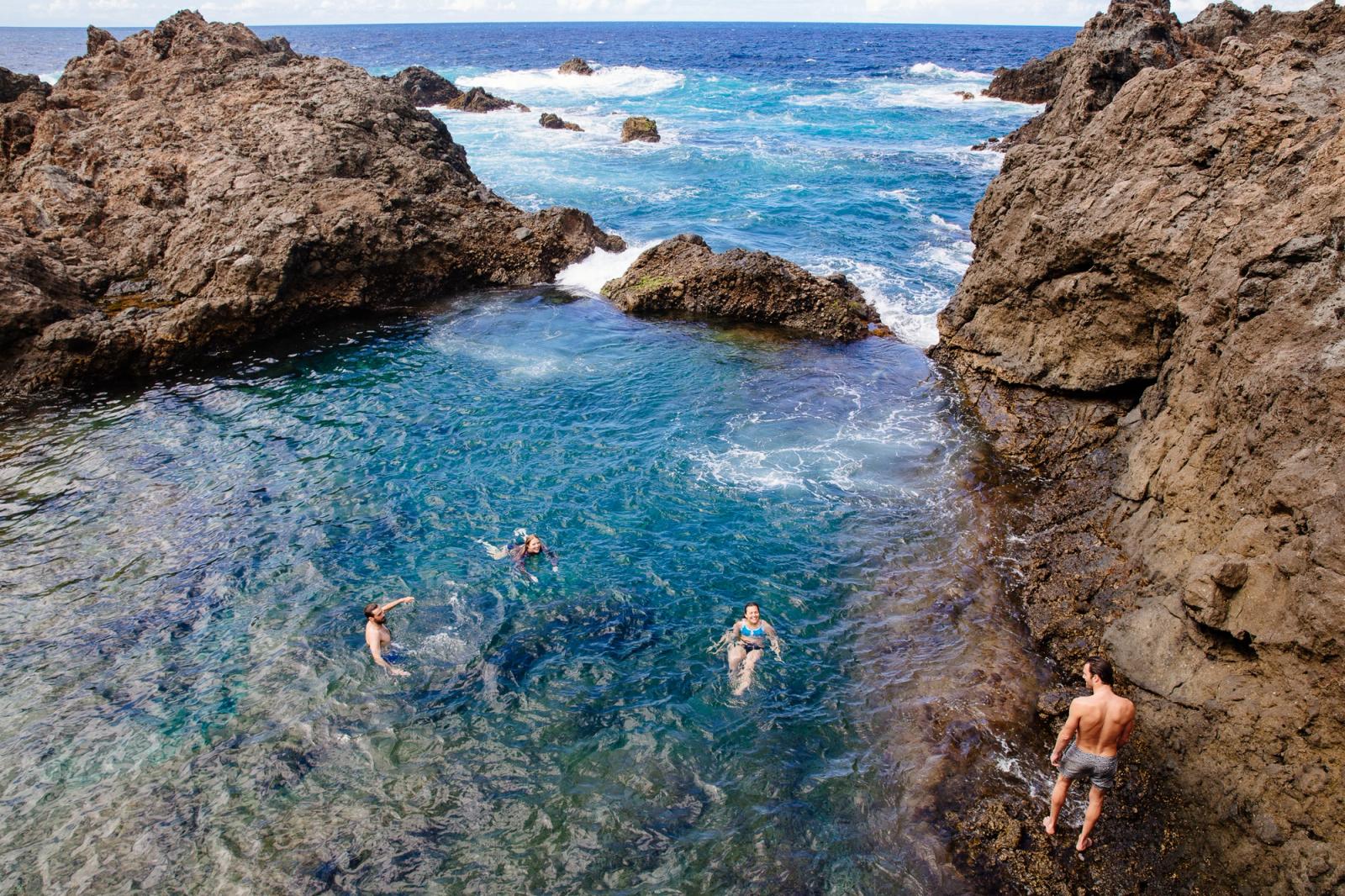 A group of people swimming in a natural rock pool surrounded by large boulders and ocean waves.