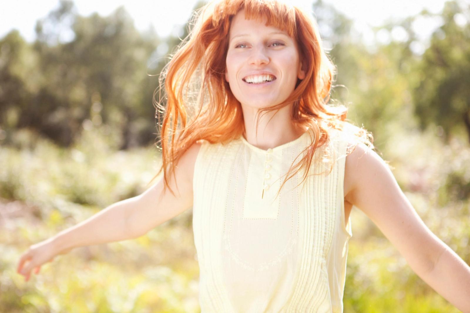 A woman with long, curly red hair and a yellow sundress is running through a field of tall grass, laughing and smiling.