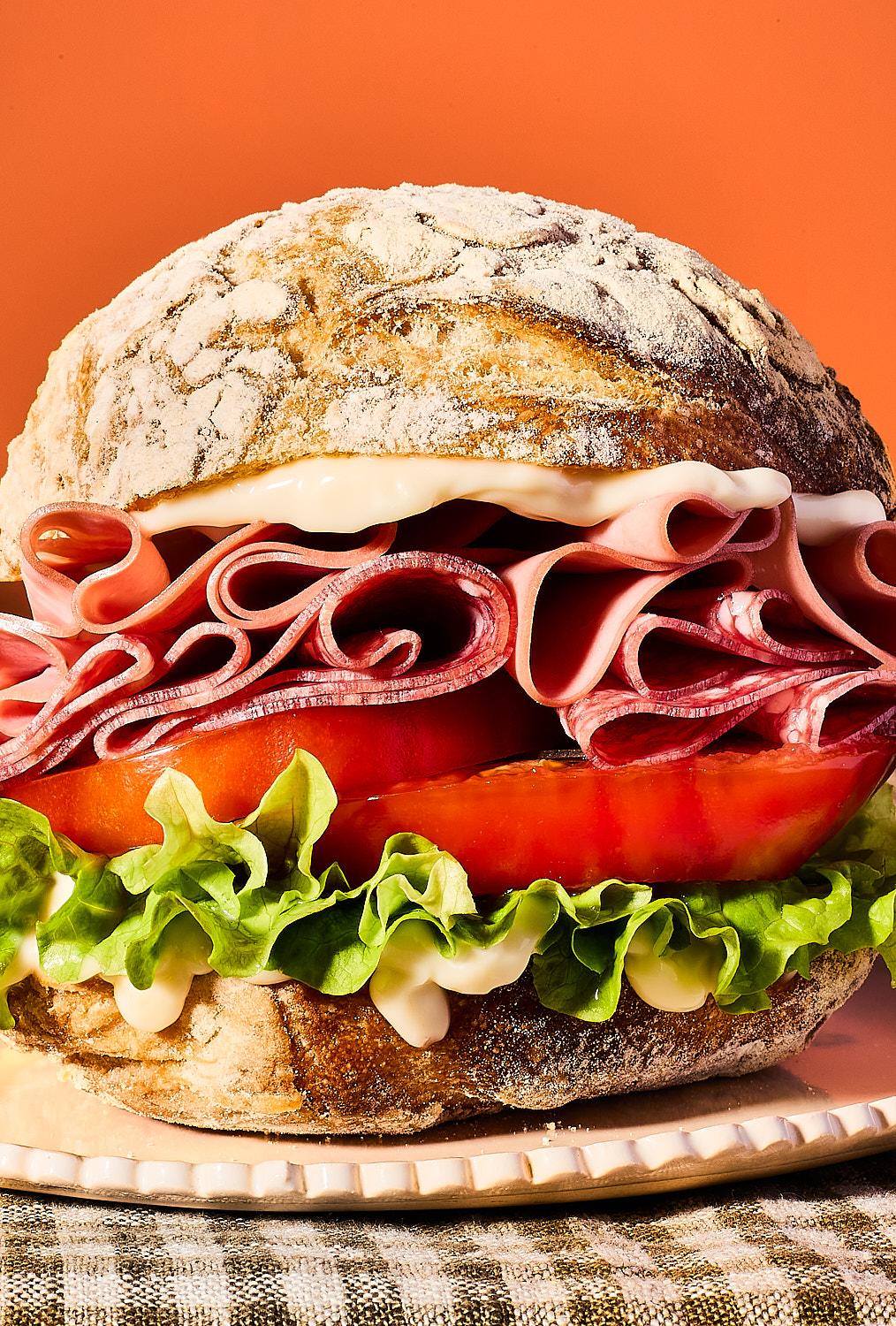 he image is a macro shot of a large sandwich on a crusty roll, showcasing thick layers of folded mortadella, salami, fresh tomato, and curly lettuce with a generous spread of mayonnaise. The vibrant colors are set against a warm orange backdrop.