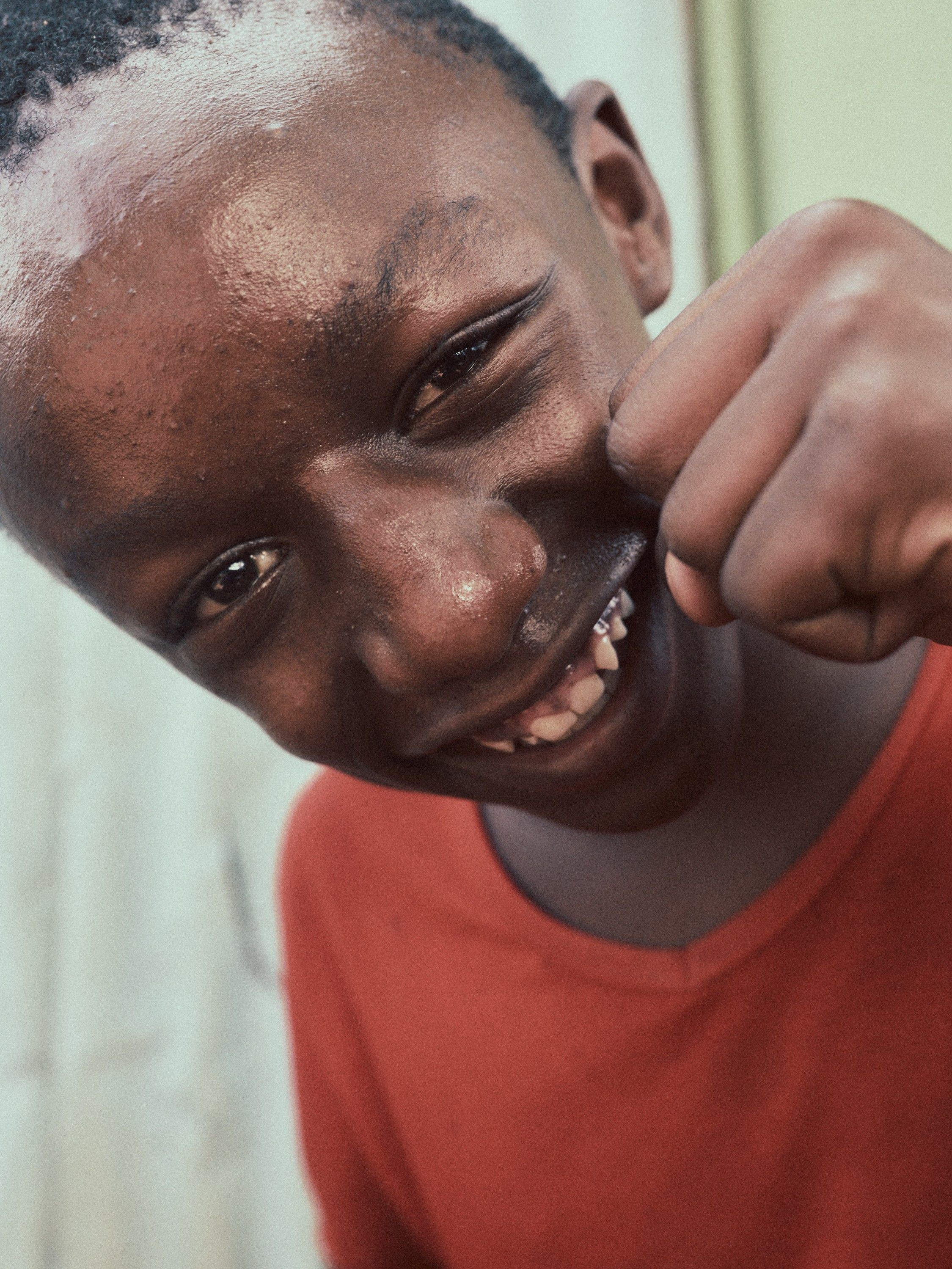 A young black boy with a mischievous grin, wearing a red t-shirt, making a 'smack your lips' gesture with his fingers.