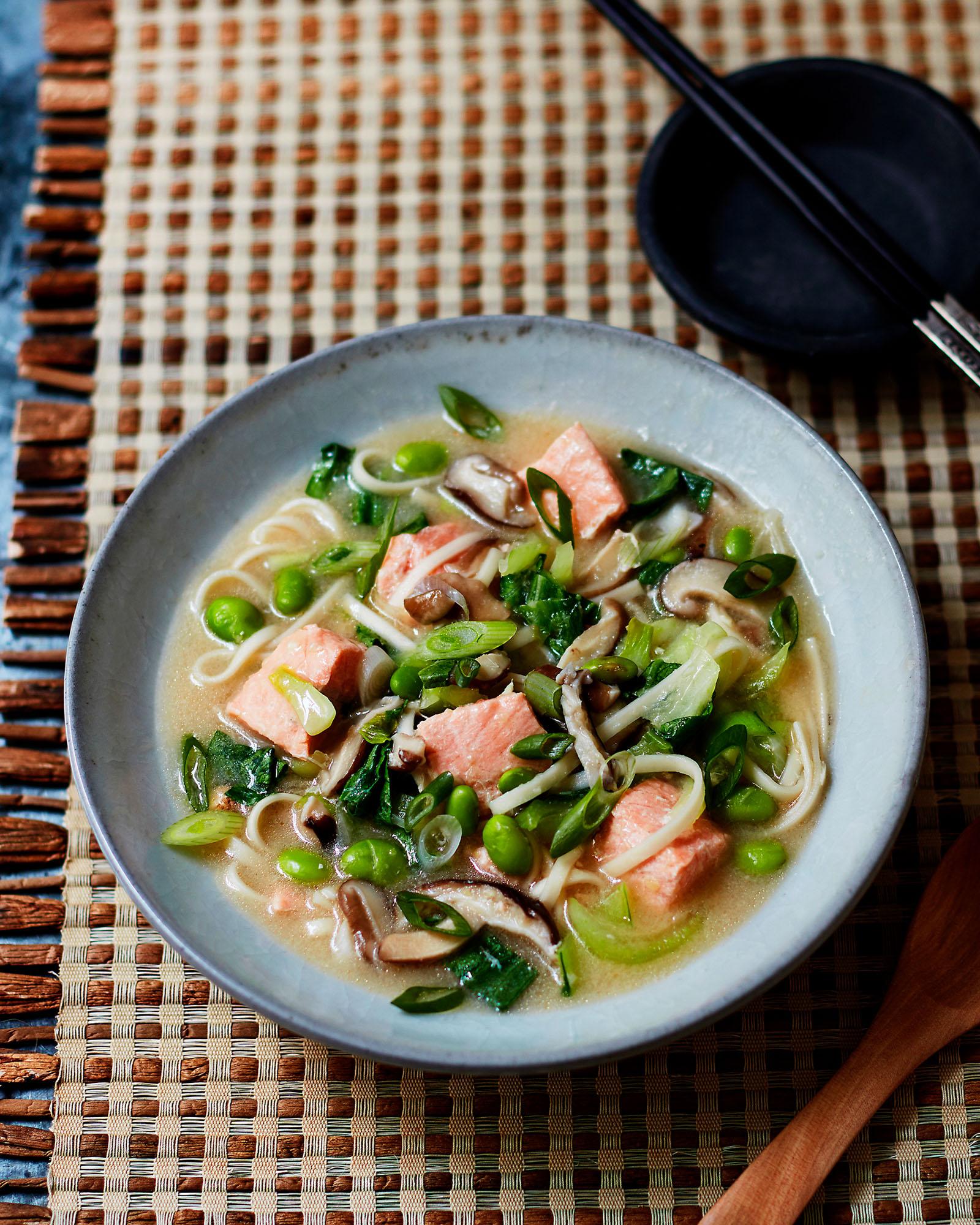 A bowl of noodle soup with salmon, vegetables, and noodles, served on a woven placemat.