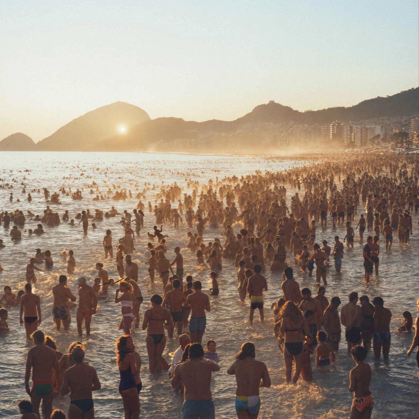 A large crowd of people in swimsuits and beachwear are gathered on a beach at sunset, with the sun setting over the ocean in the background.