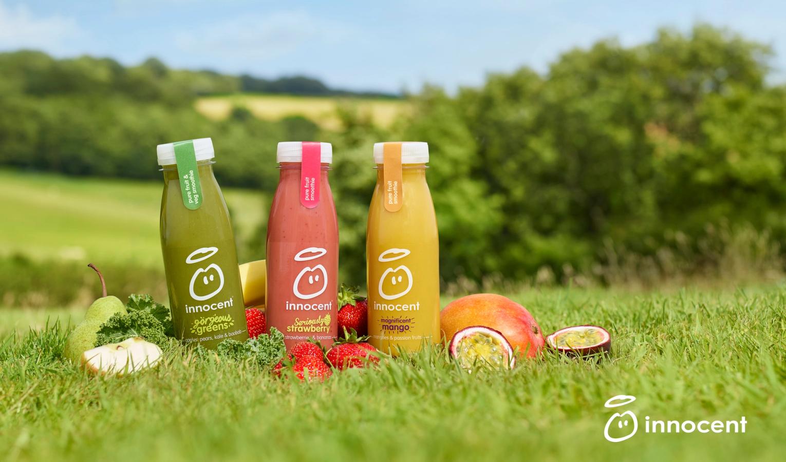 Three bottles of juice, one green, one red, and one yellow, surrounded by fruit and vegetables in a grassy field.