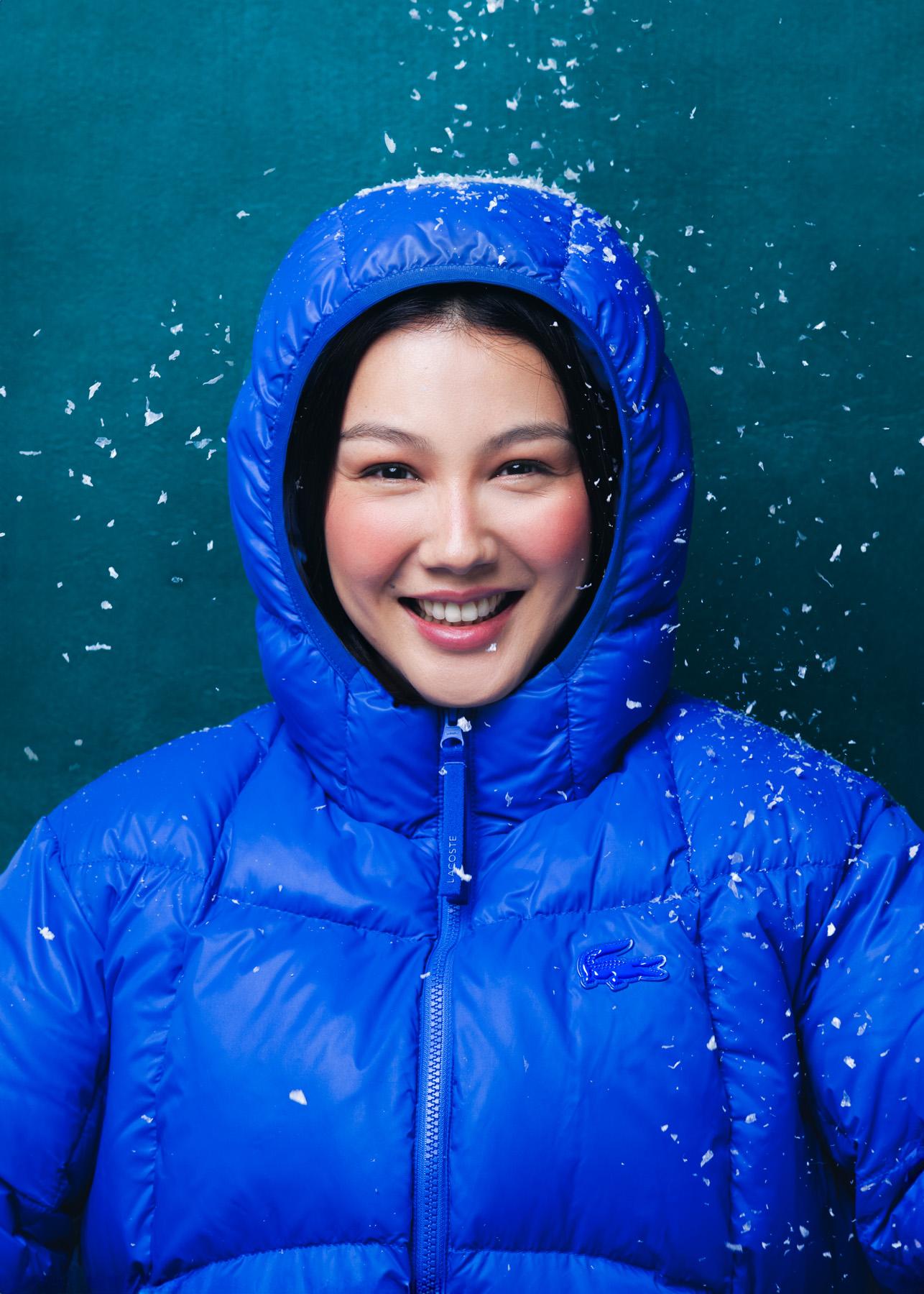 A woman in a blue puffy jacket and snow-covered hat, smiling and looking directly at the camera, with snowflakes falling around her.