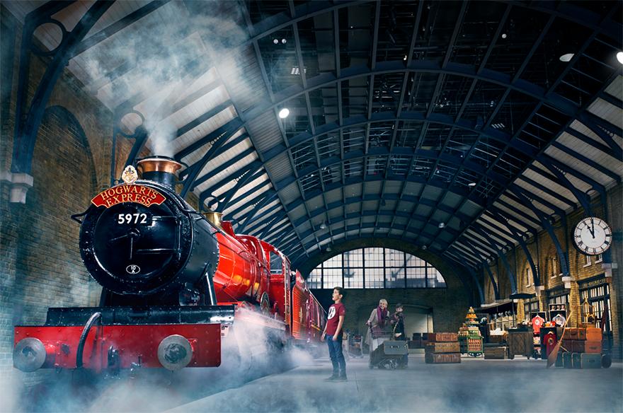 A steam train named Hogwarts Express is in a large, dimly lit Victorian train station with a clock and sign, and some people are visible in the background.