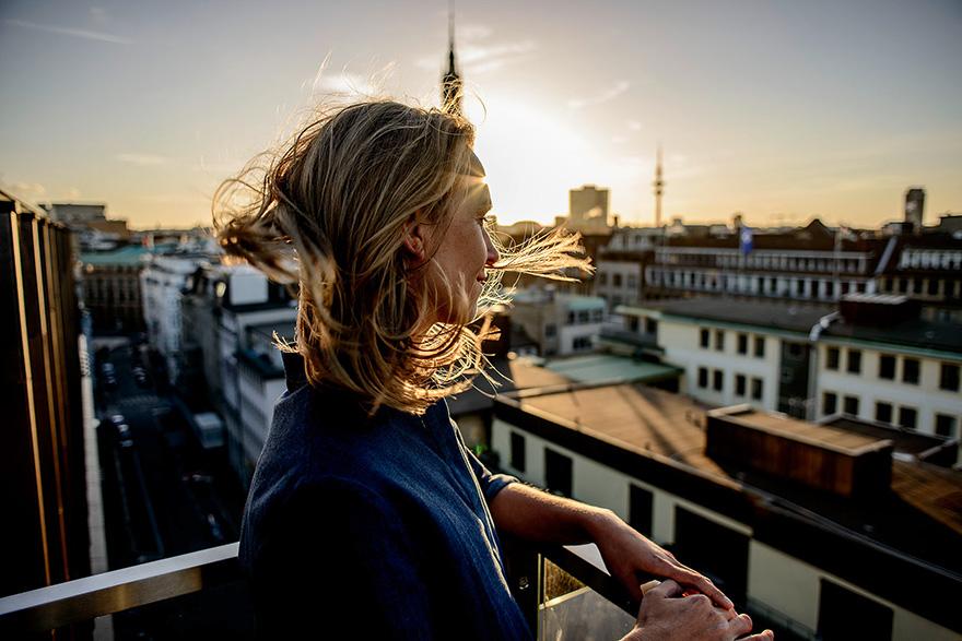 A woman with long blonde hair stands on a rooftop, looking out over a city skyline at sunset.